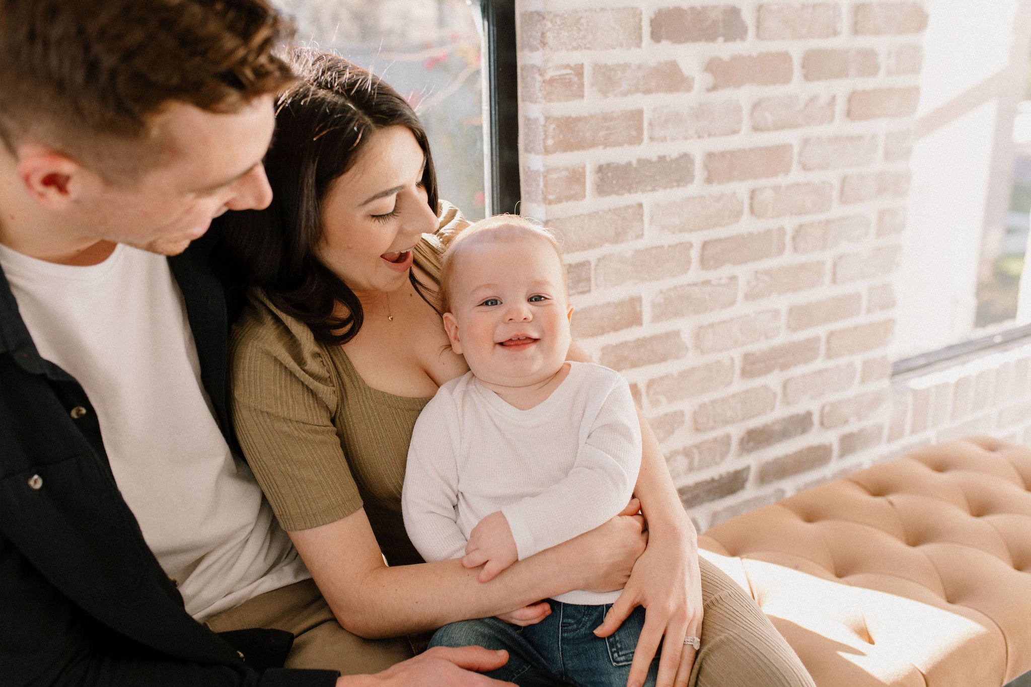 baby boy smiling with mom and dad in neutral studio space with red brick wall in Phoenix, Arizona