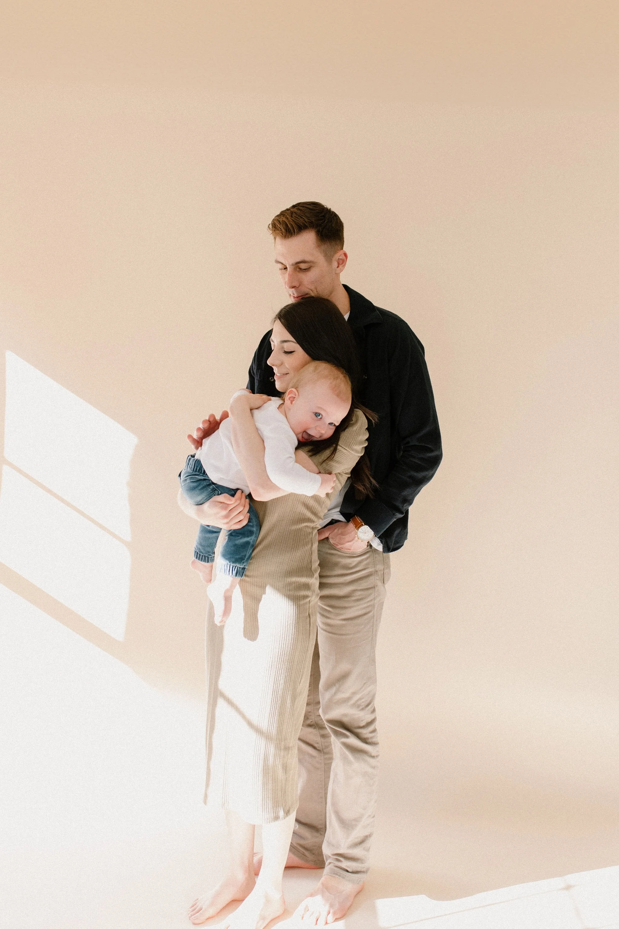 mom holding baby boy with dad hugging them both in neutral studio space with beautiful window light in Phoenix, arIzonA