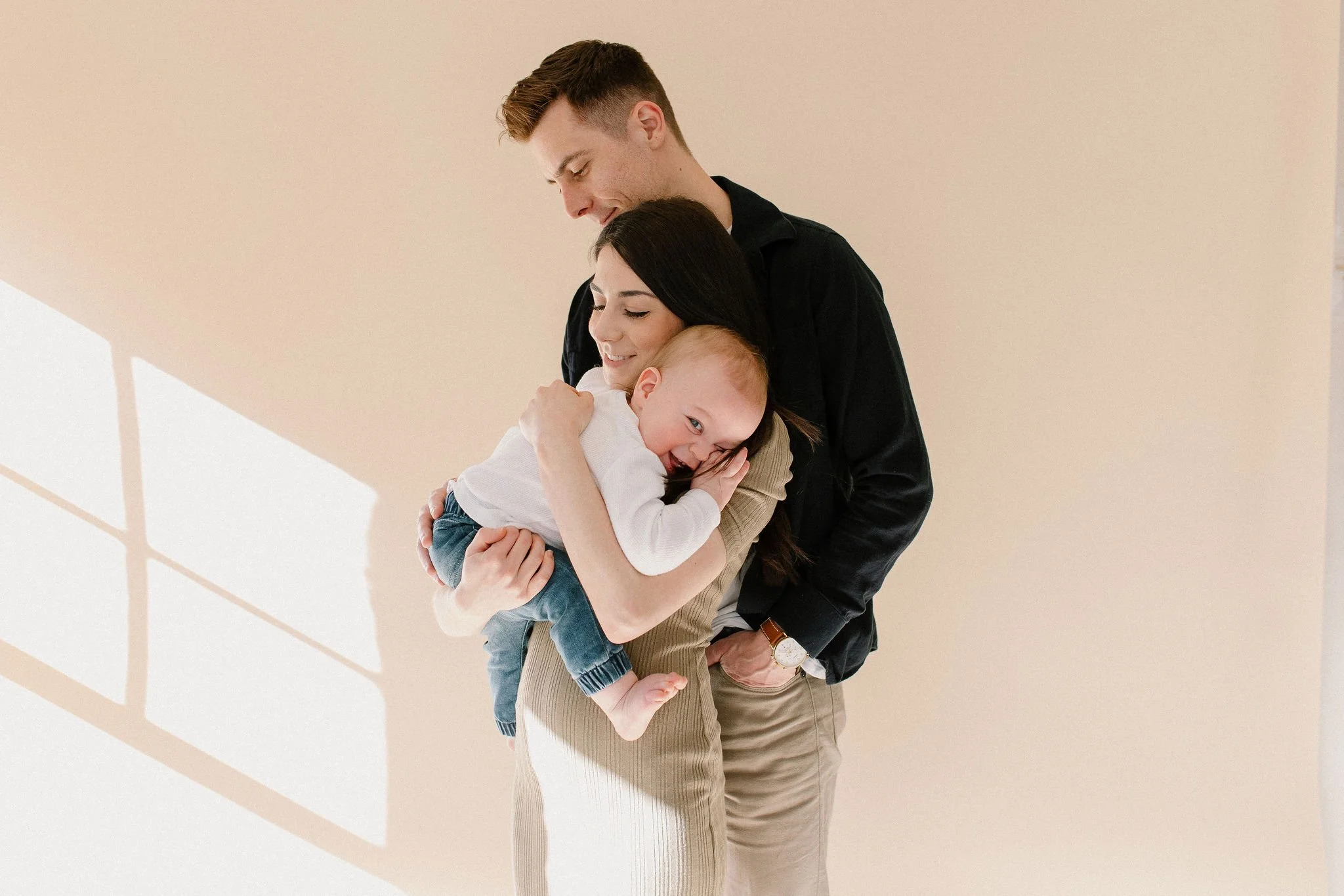 baby boy hugging mom and smiling while dad hugs his family at neutral studio in Phoenix, Arizona