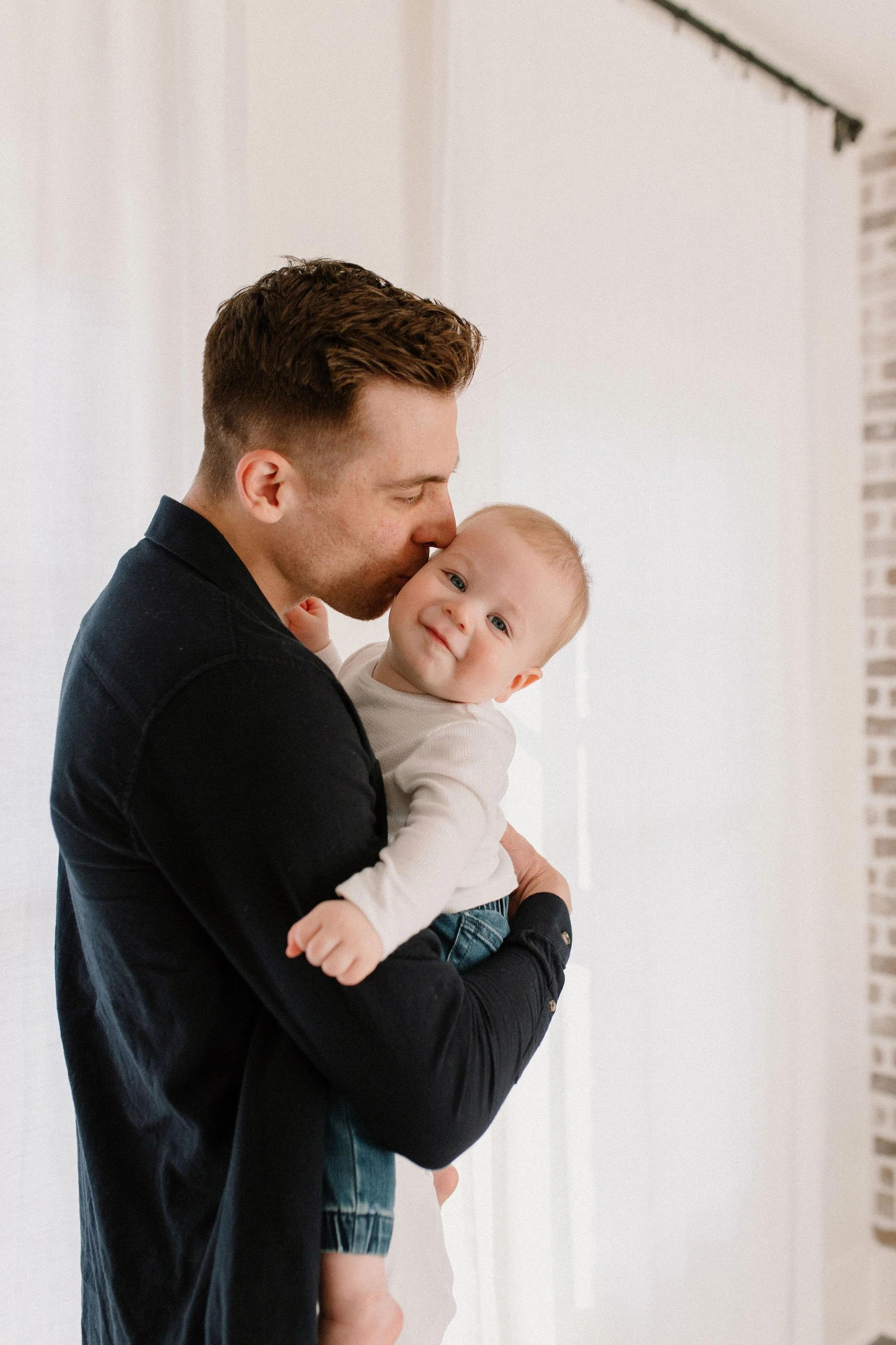 baby boy smiling while dad kisses his cheek during family photo session at neutral studio in Phoenix, Arizona