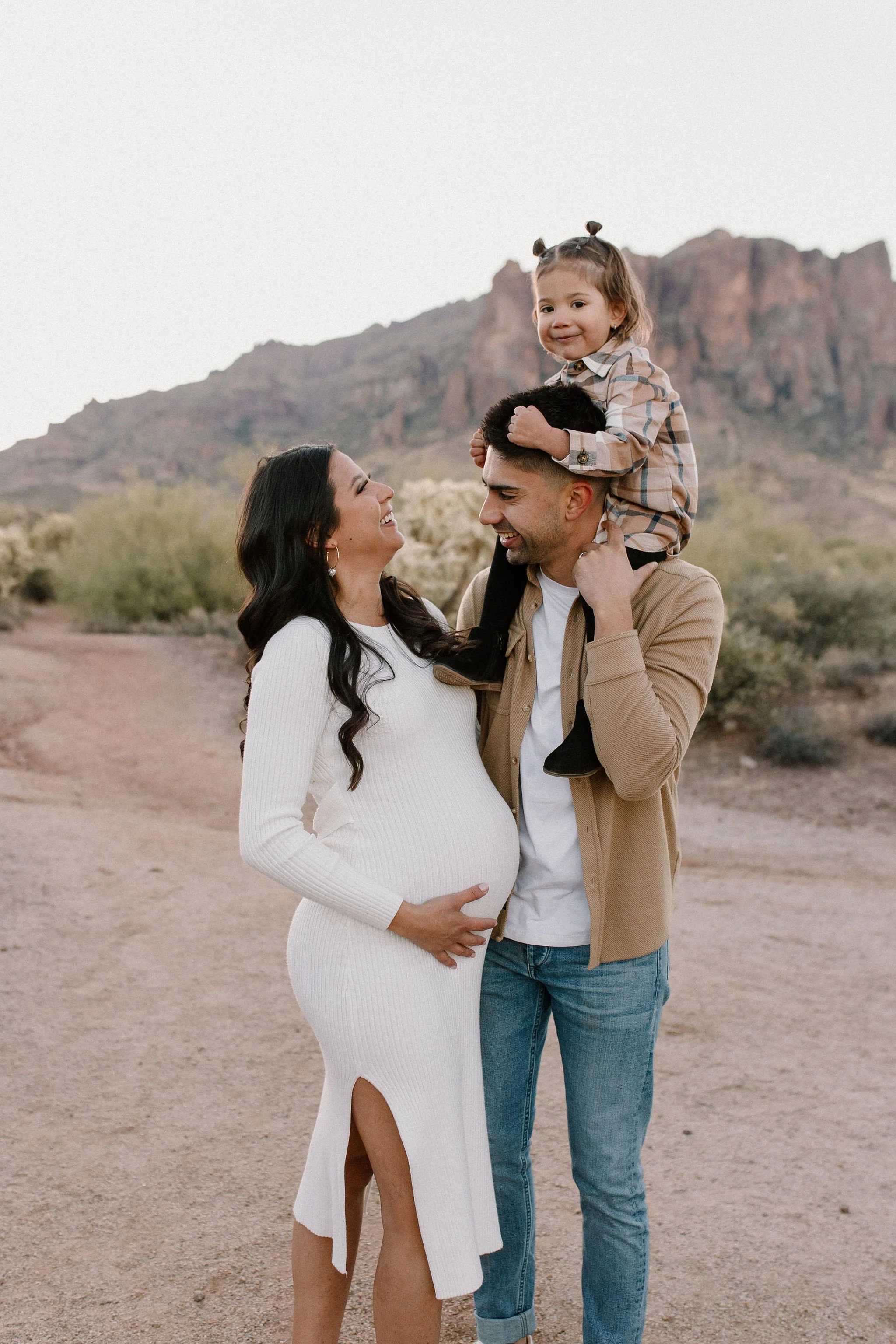 pregnant mom holding belly hugging dad with toddler on his shoulders in desert family maternity pics at Lost Dutchman State Park
