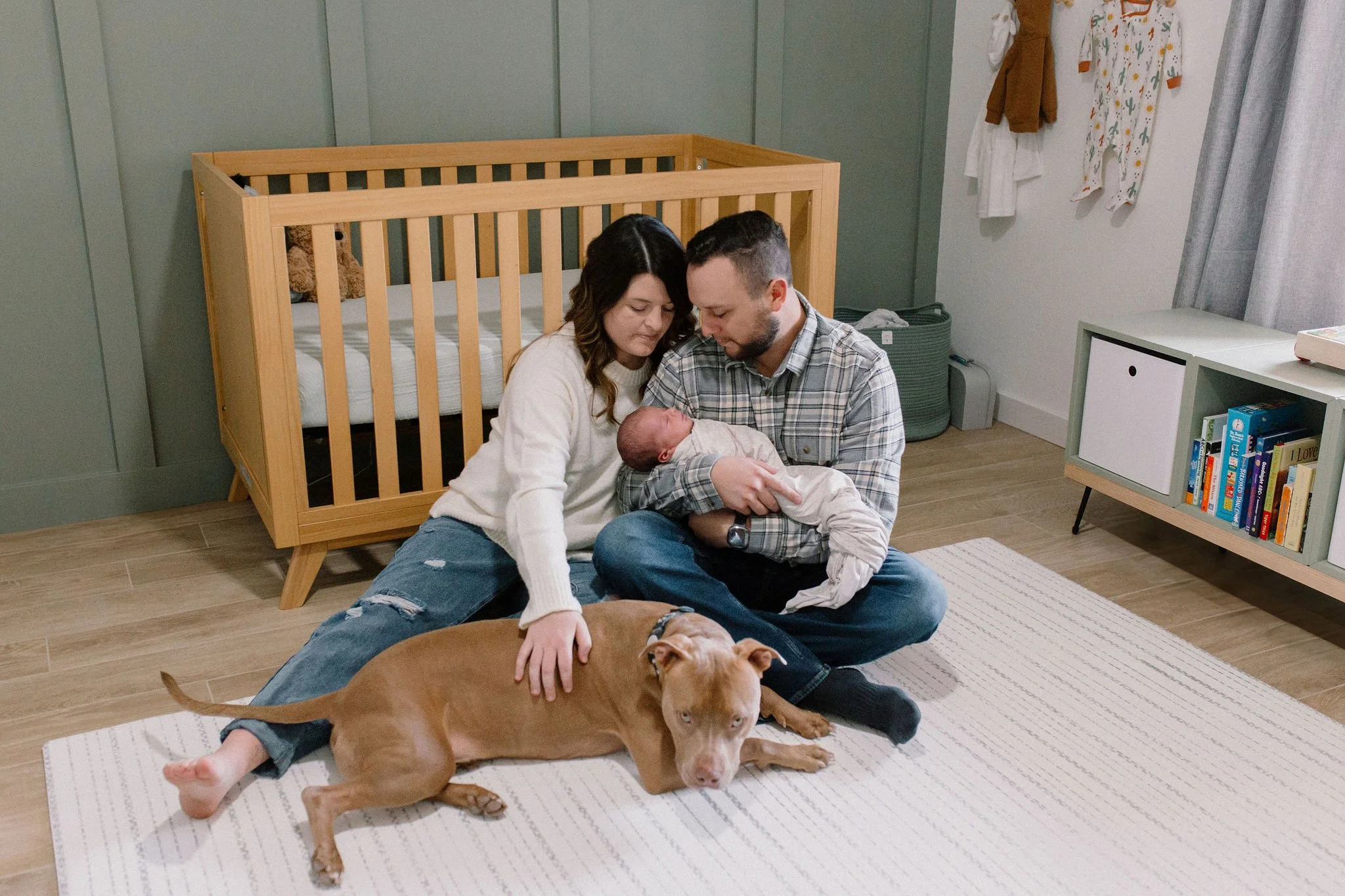 family sitting next to crib holding newborn baby in neutral green nursery with dog