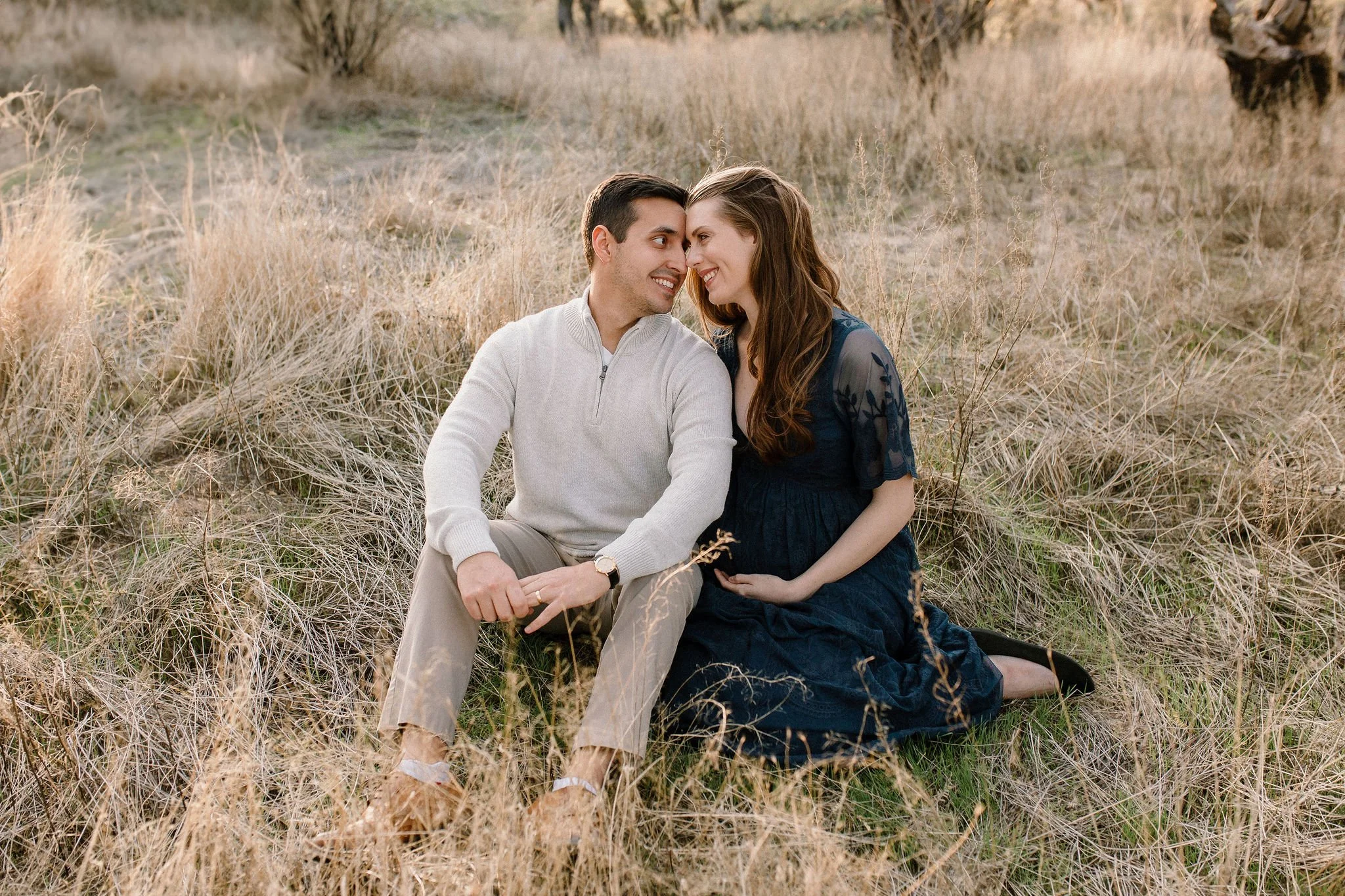 pregnant mom sitting with husband looking at each other in tall yellow grass during Mesa Arizona maternity photography session