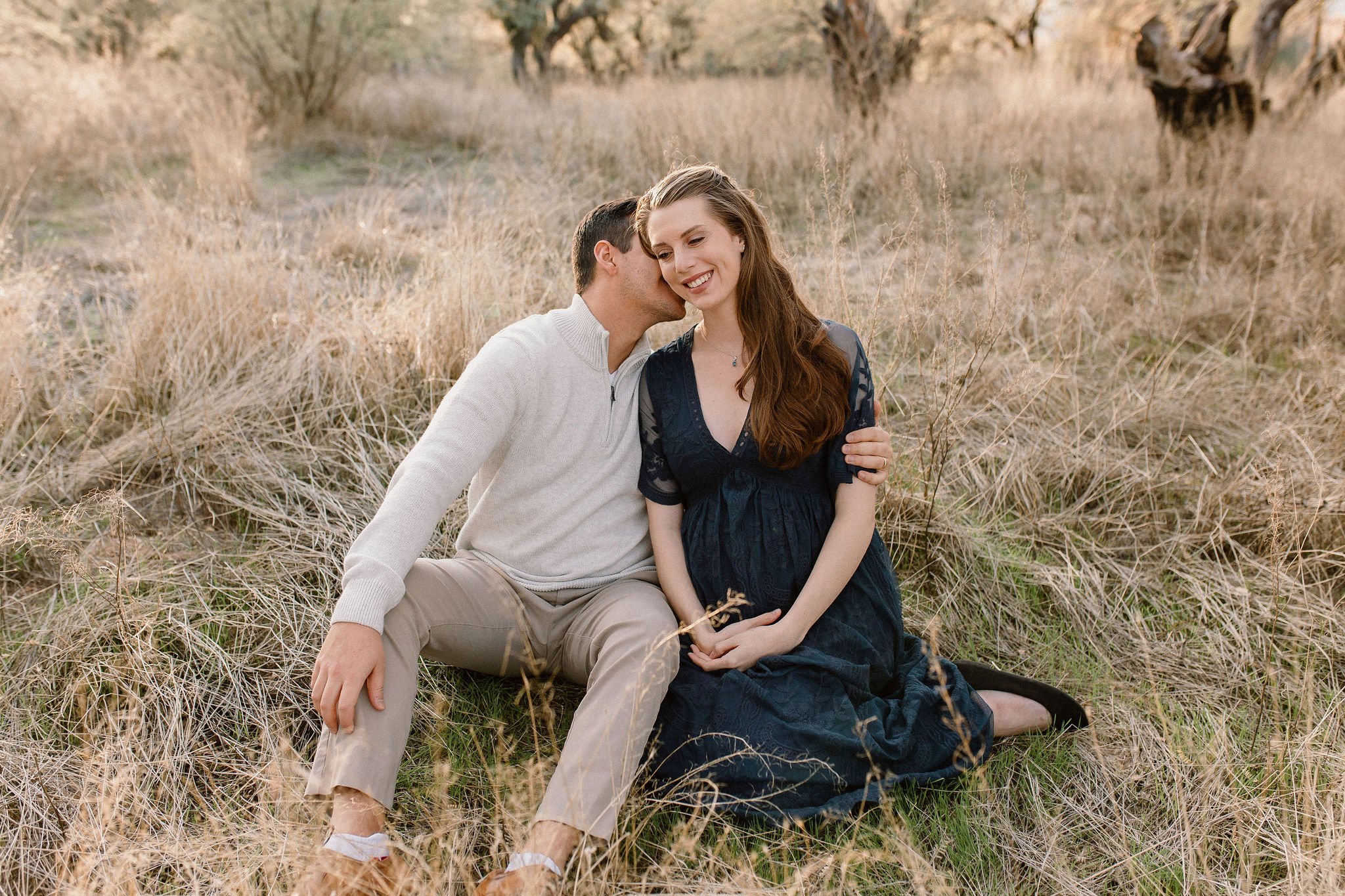 pregnant mom sitting with husband kissing her cheek in tall yellow grass during Mesa maternity photography session