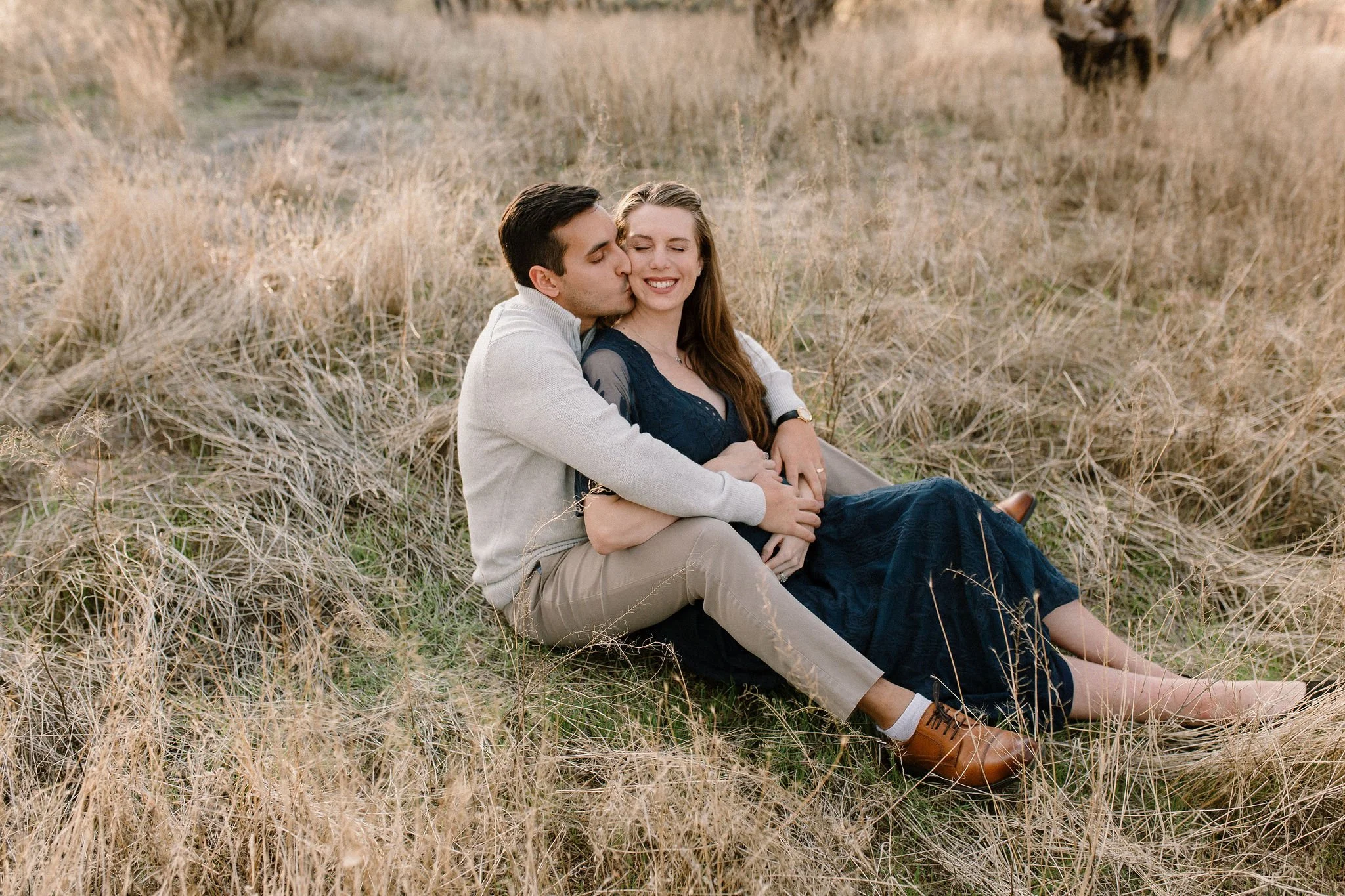 pregnant mom sitting with husband kissing her cheek in tall yellow grass during Mesa maternity photography session