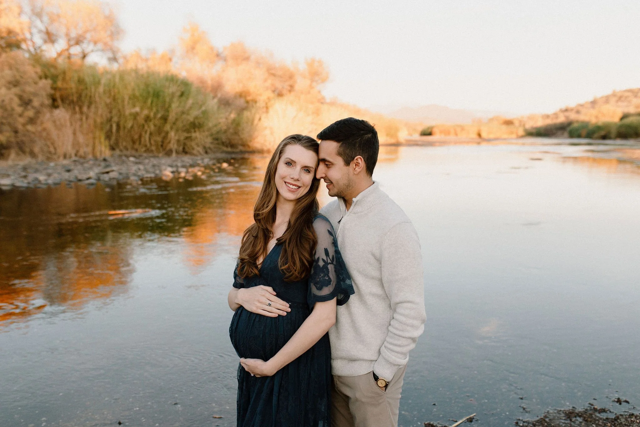 pregnant mom holding belly and husband looking at her by Salt River in Mesa maternity photography session
