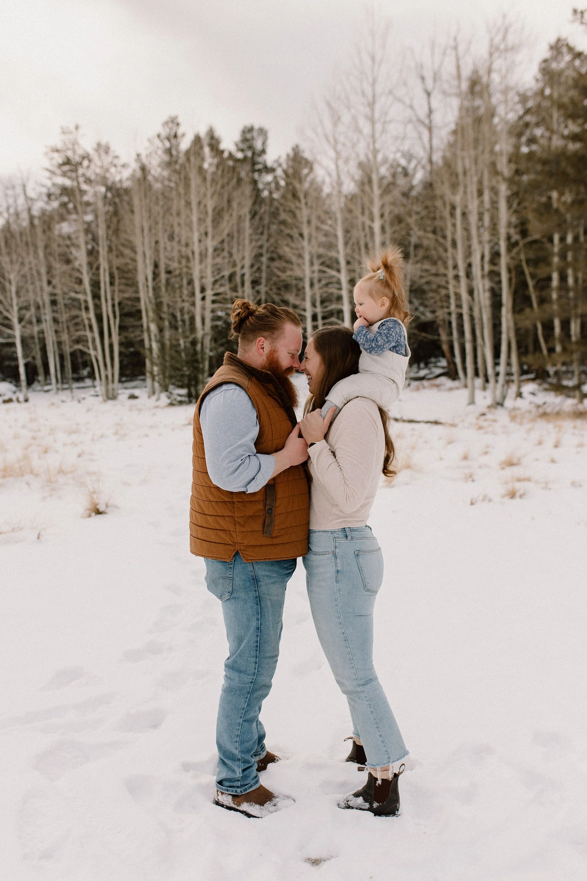 family of three standing together mom and dad close together looking at each other with noses touching and baby girl on mom's shoulders in snowy family photography session in Flagstaff Arizona
