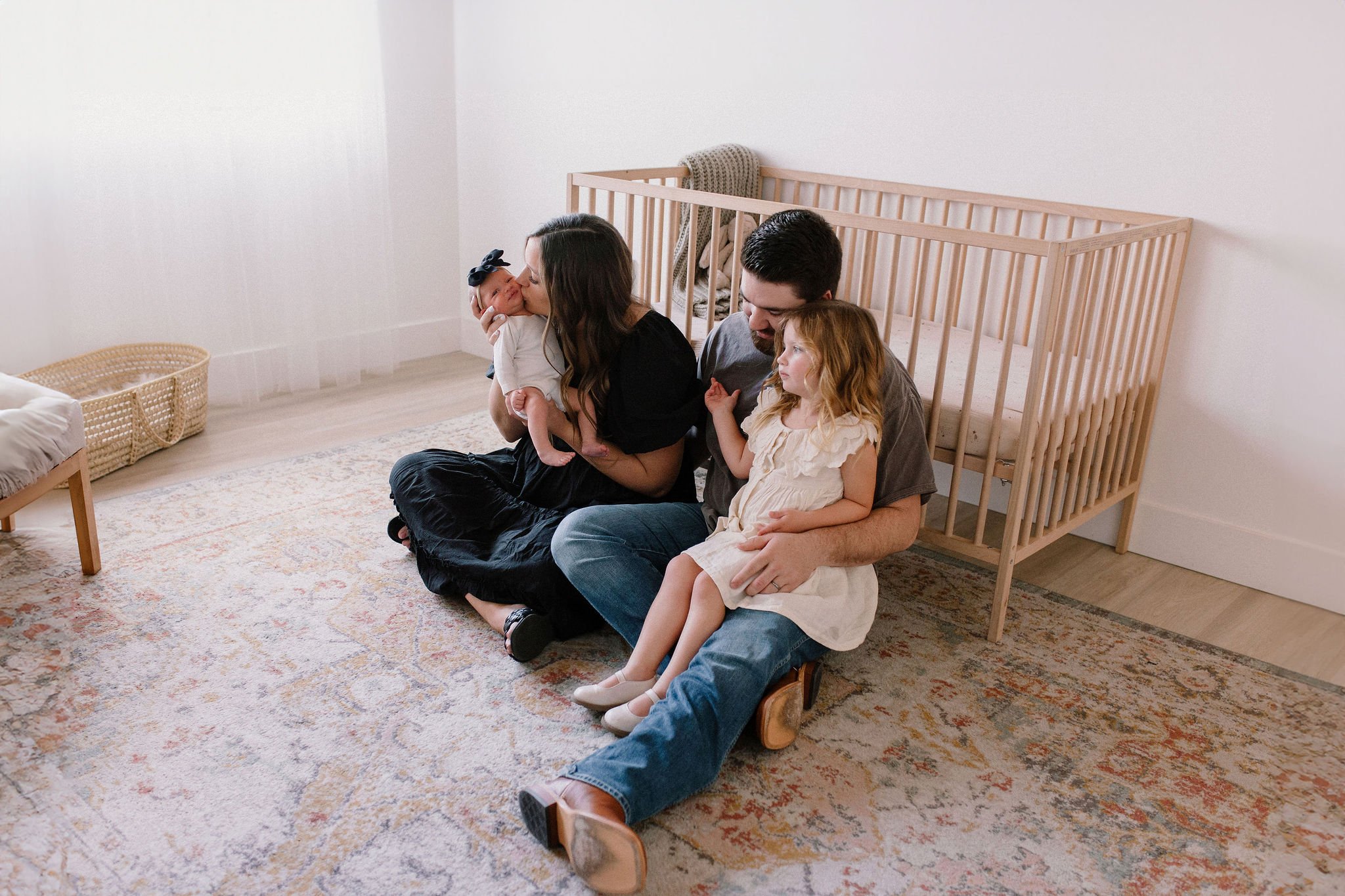 family sitting together next to crib with dad holding toddler and mom kissing newborn baby in neutral nursery