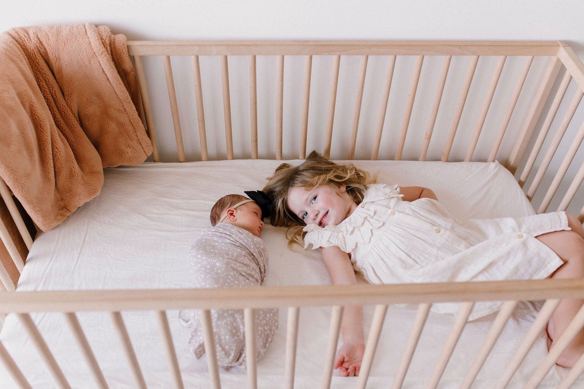 big sister laying in crib with swaddled newborn baby sister in neutral studio