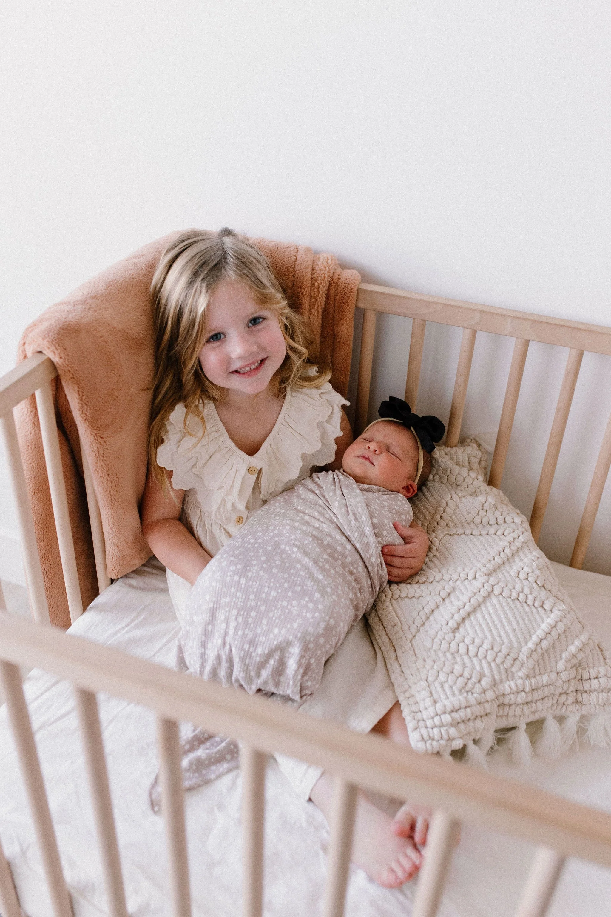 big sister holding swaddled newborn baby sister in crib in neutral nursery