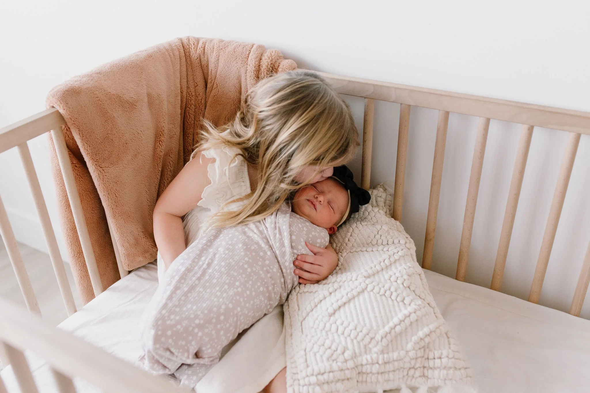 big sister holding and kissing swaddled baby sister in crib in neutral nursery