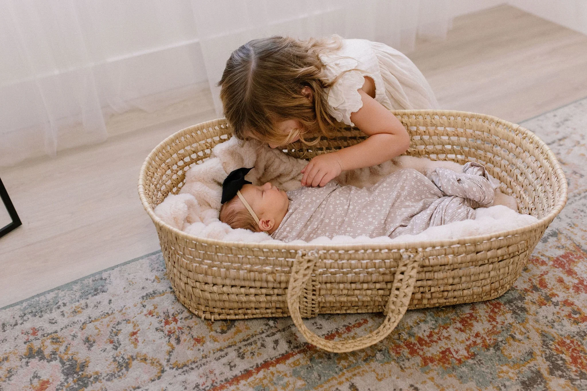 big sister looking in at swaddled newborn baby sister in Moses basket in neutral nursery
