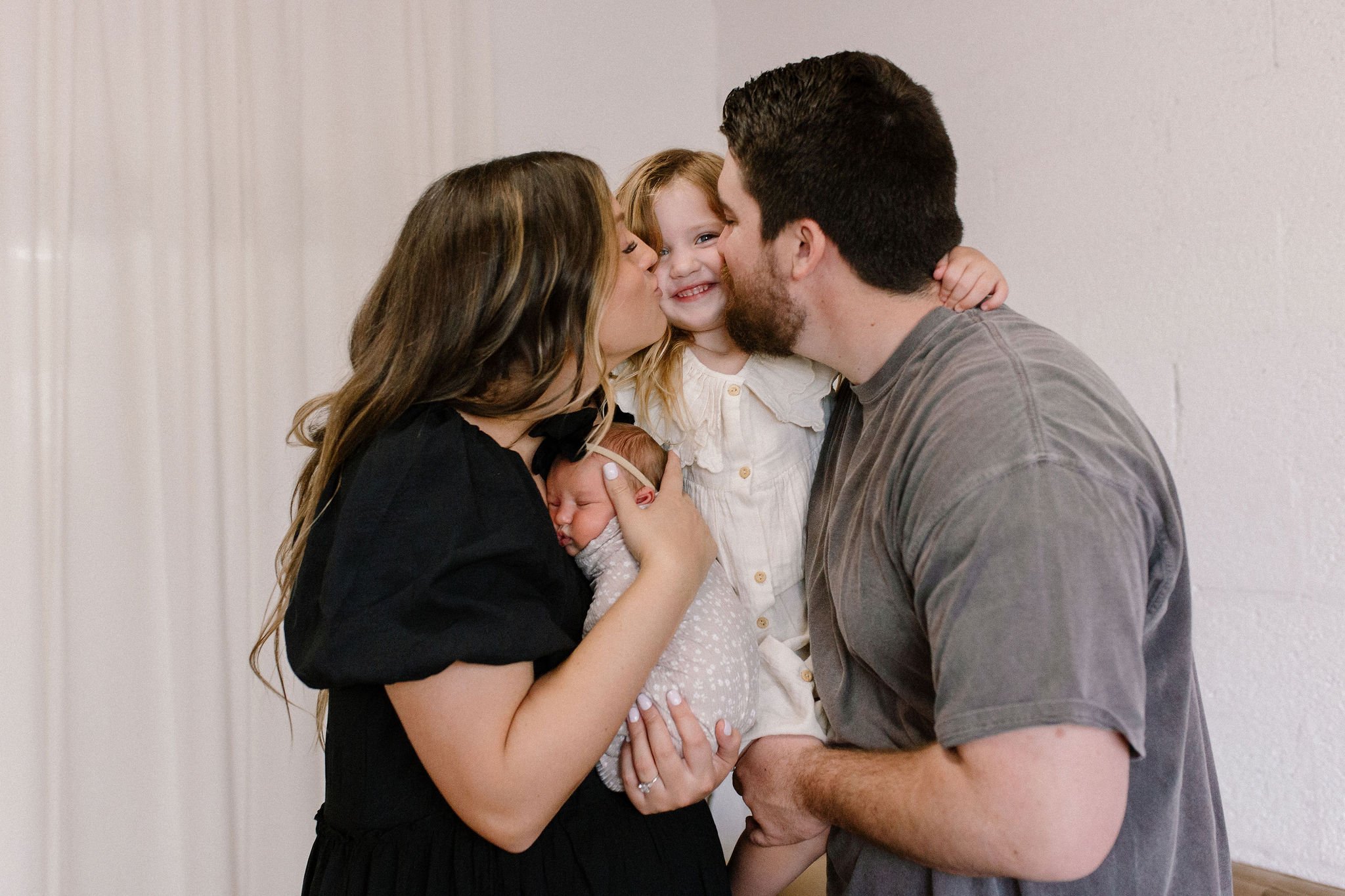 mom and dad holding newborn baby and kissing toddler daughter's cheek in neutral nursery