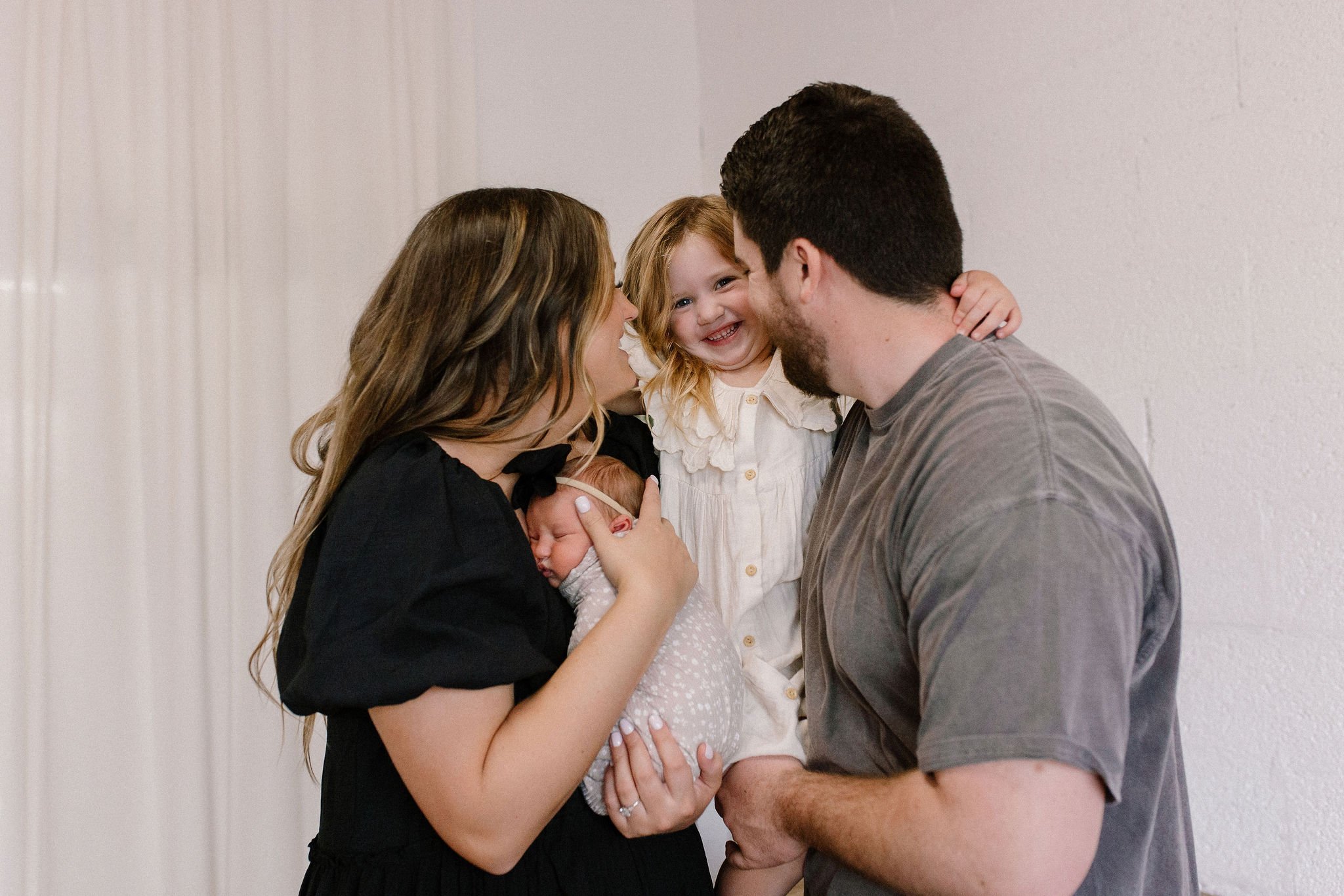 mom and dad holding newborn baby and smiling at toddler in neutral nursery