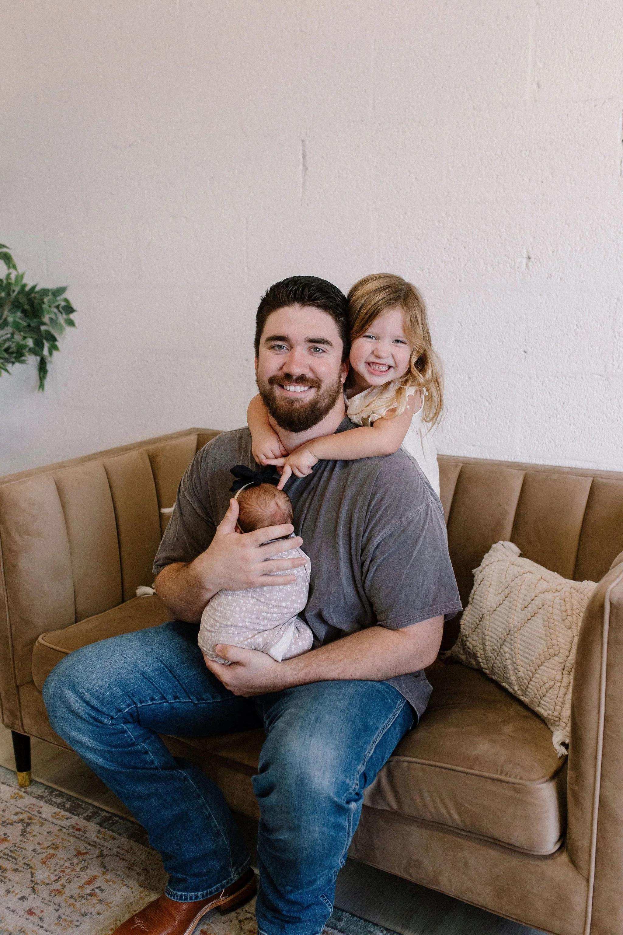 dad with his girls newborn baby and toddler sitting close on couch in neutral nursery