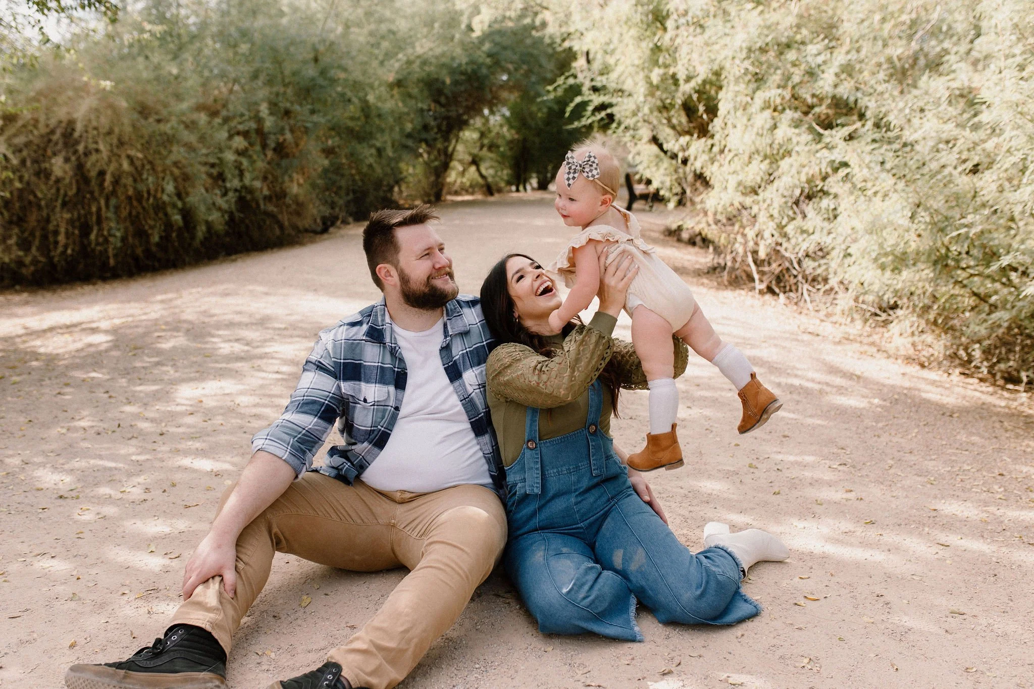 family of three sitting on the ground in desert greenery with mom holding baby girl in the air and smiling