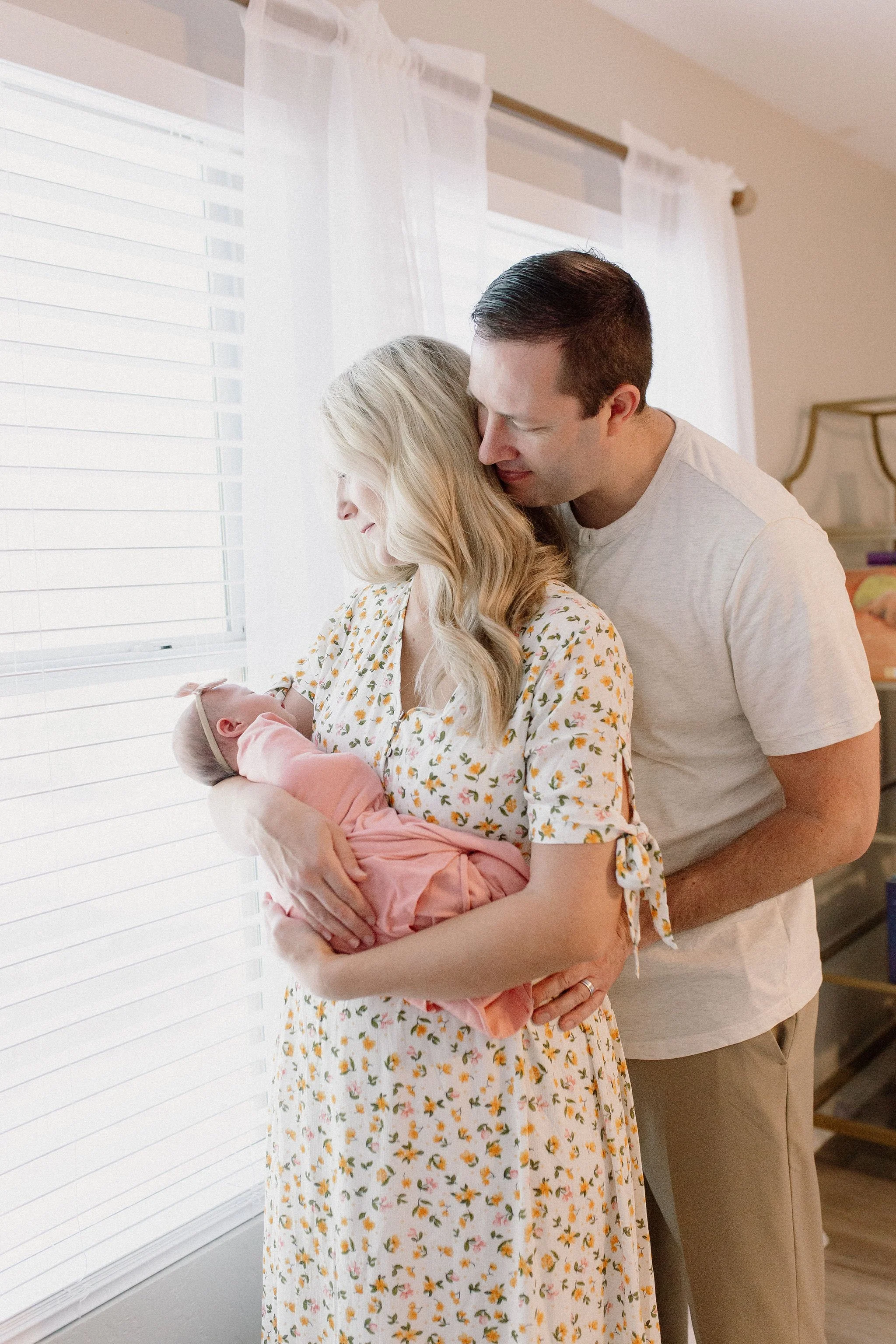 mom and dad holding swaddled newborn baby girl next to window with light shining in