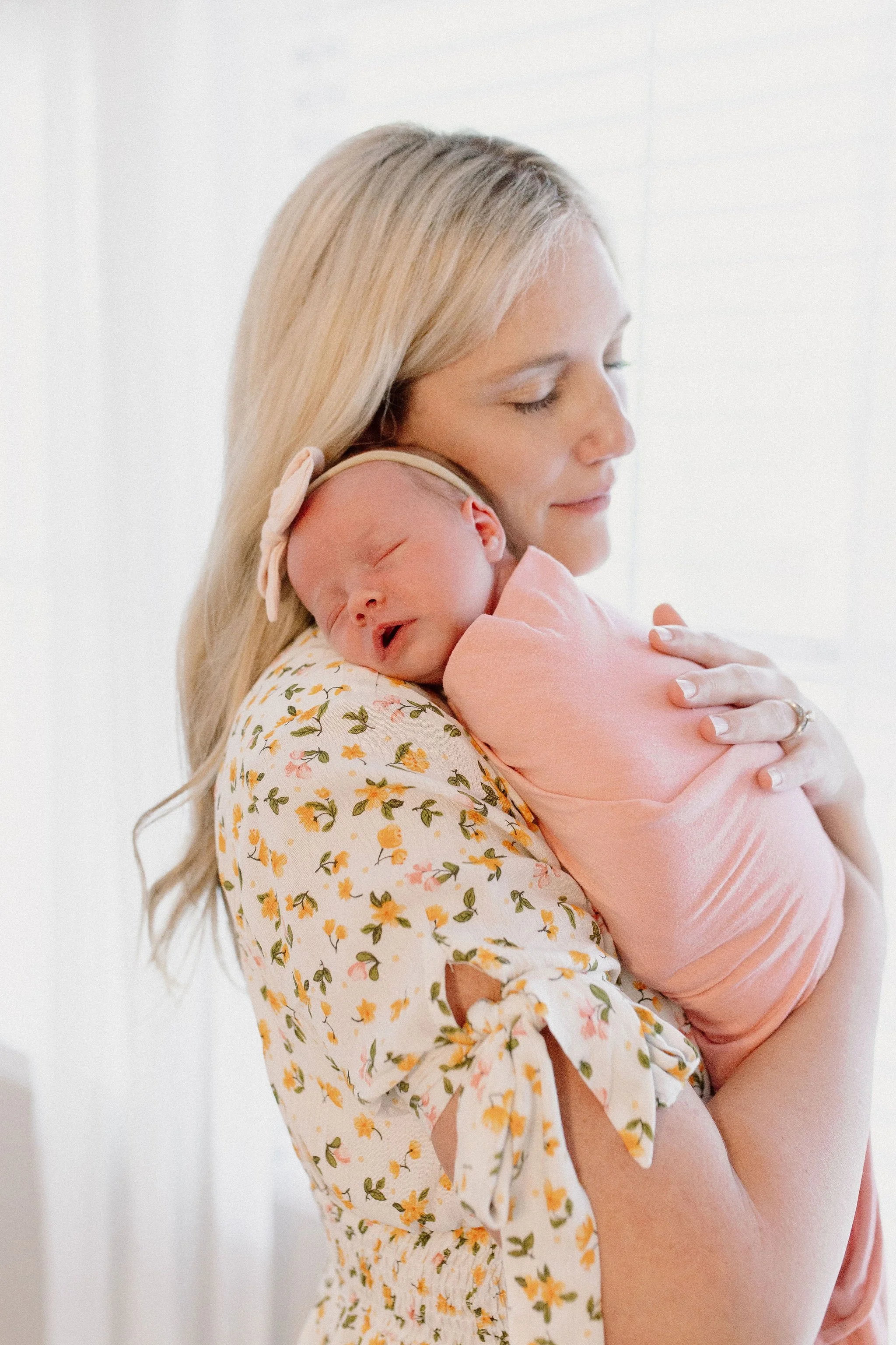 mom holding swaddled newborn baby girl in window light during lifestyle in-home photography session
