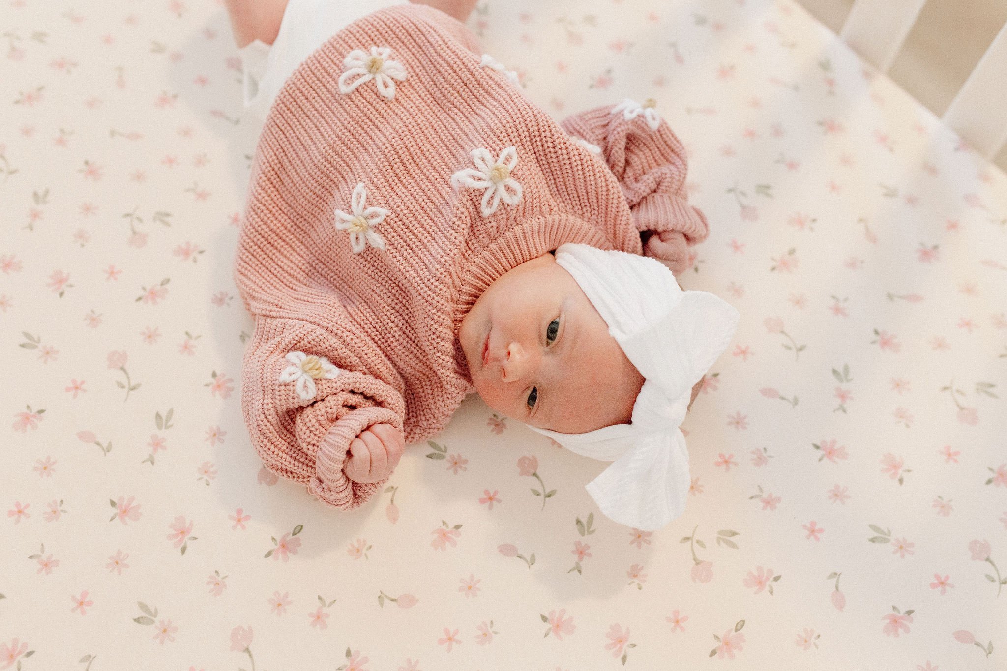 newborn baby girl in knitted floral sweater in crib during lifestyle in-home photography session