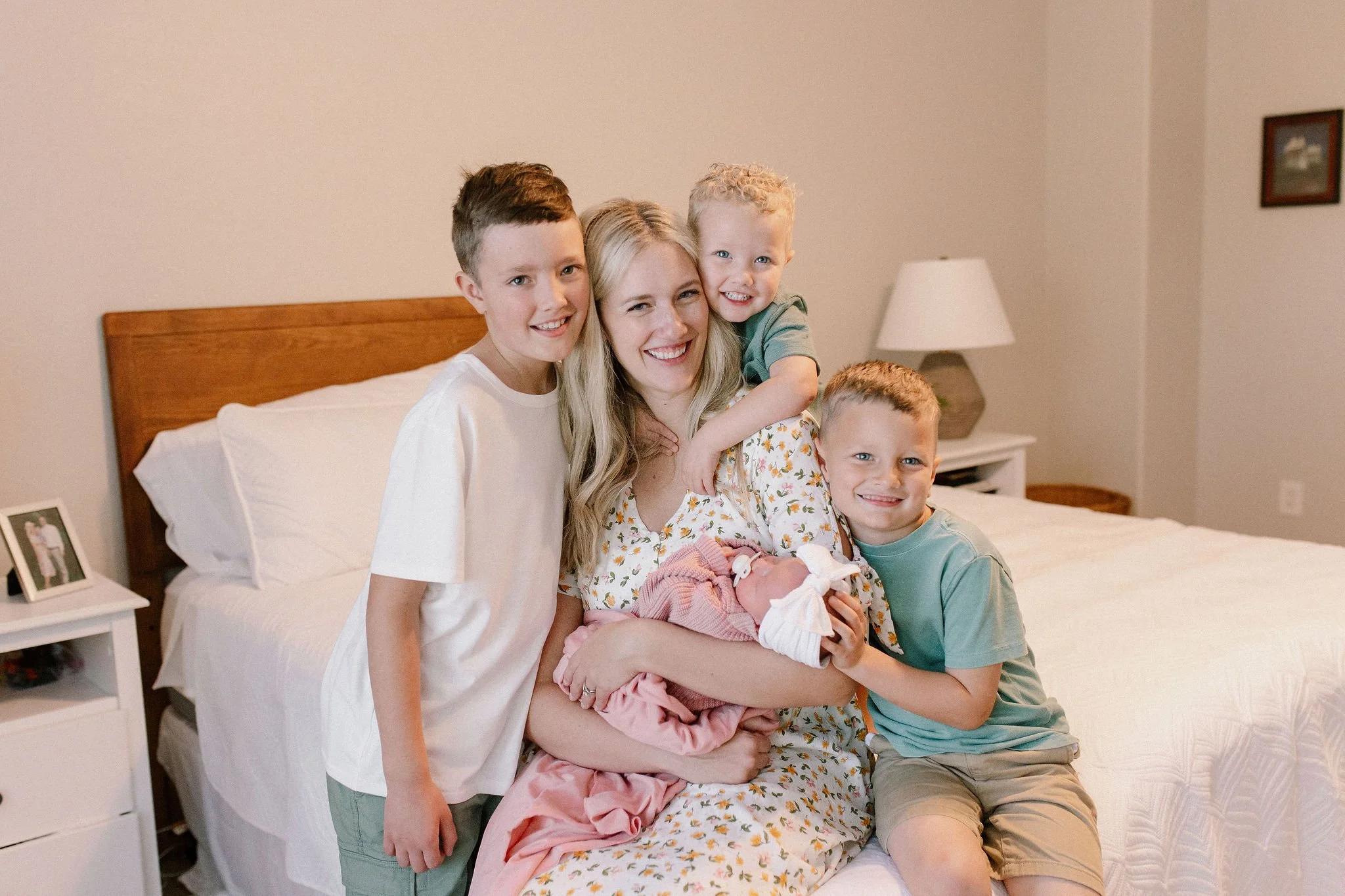 mom holding swaddled newborn baby girl with her three older brothers hugging mom in lifestyle in-home newborn photography session