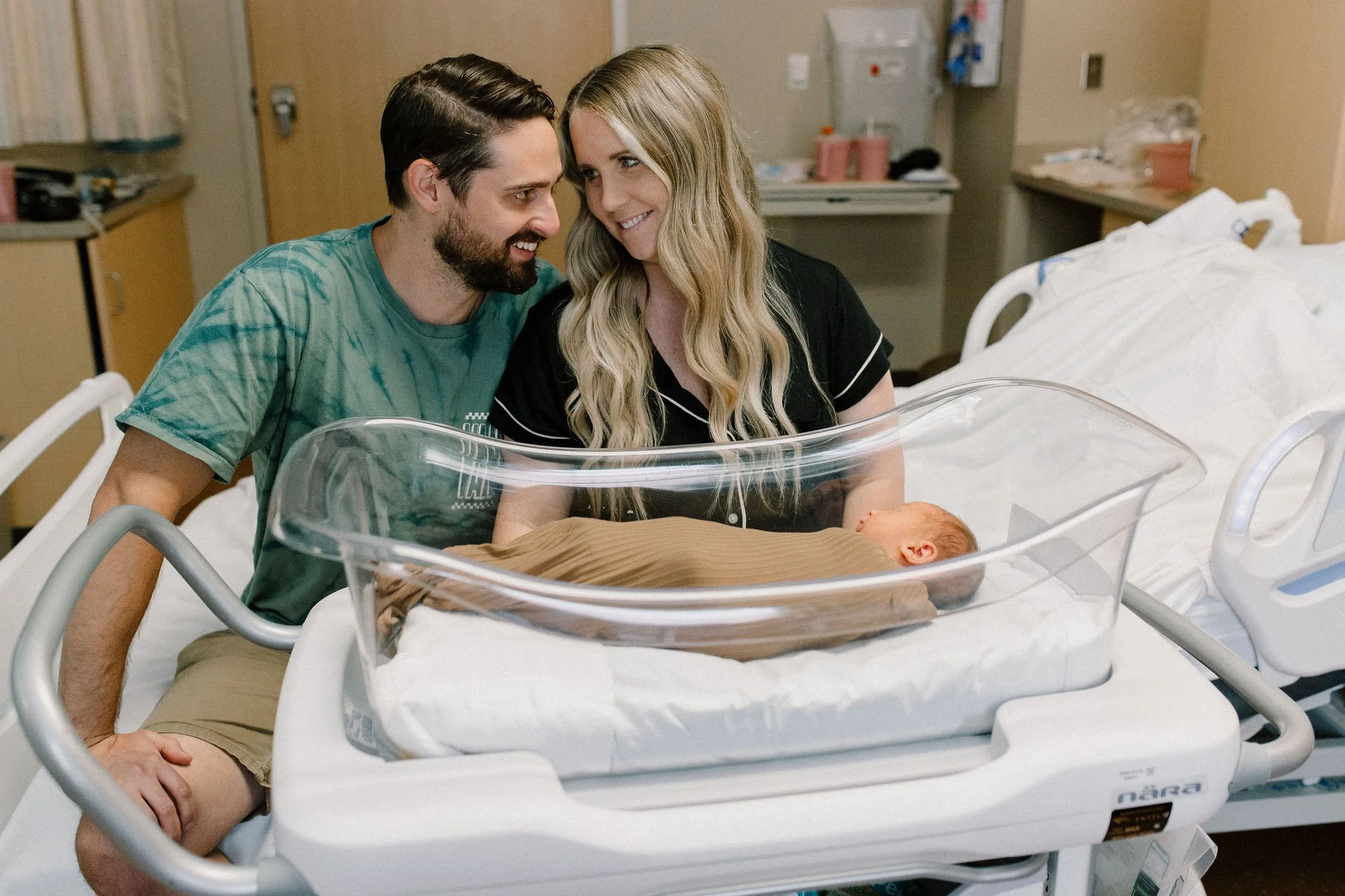 mom and dad looking at each other with newborn baby boy sleeping in hospital bassinet