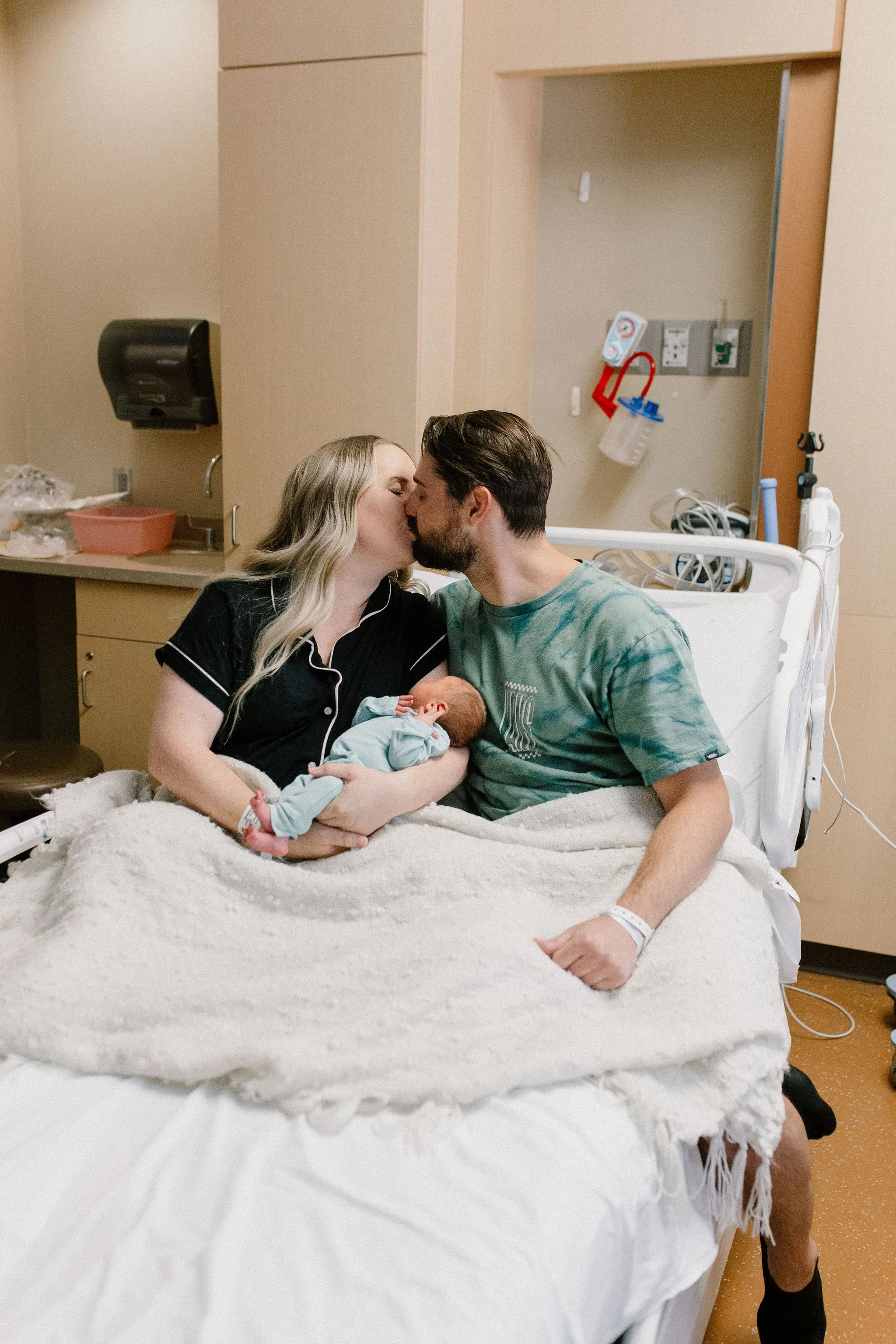 mom and dad kissing while holding newborn baby boy in hospital bed after birth