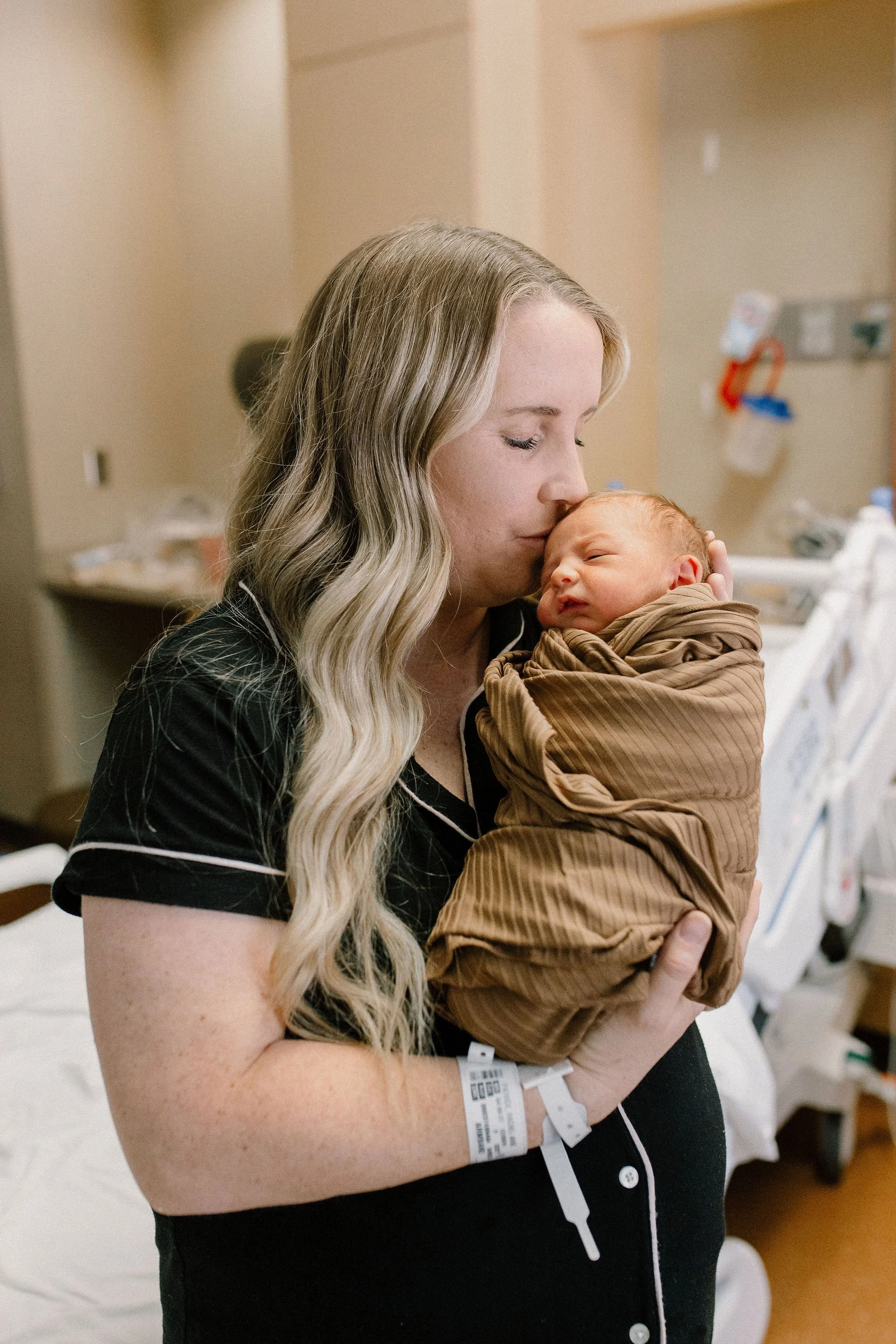 mom holding and kissing swaddled newborn baby boy in hospital room after birth