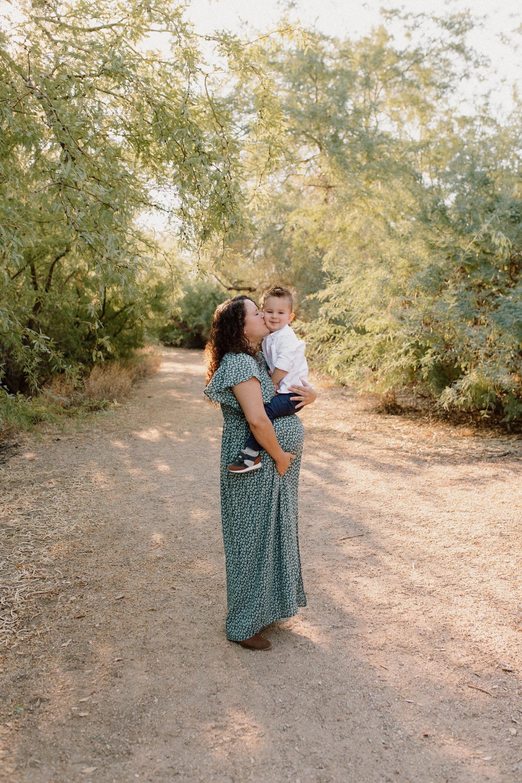 toddler sitting on pregnant mom's belly while mom kisses his face during family maternity photography session