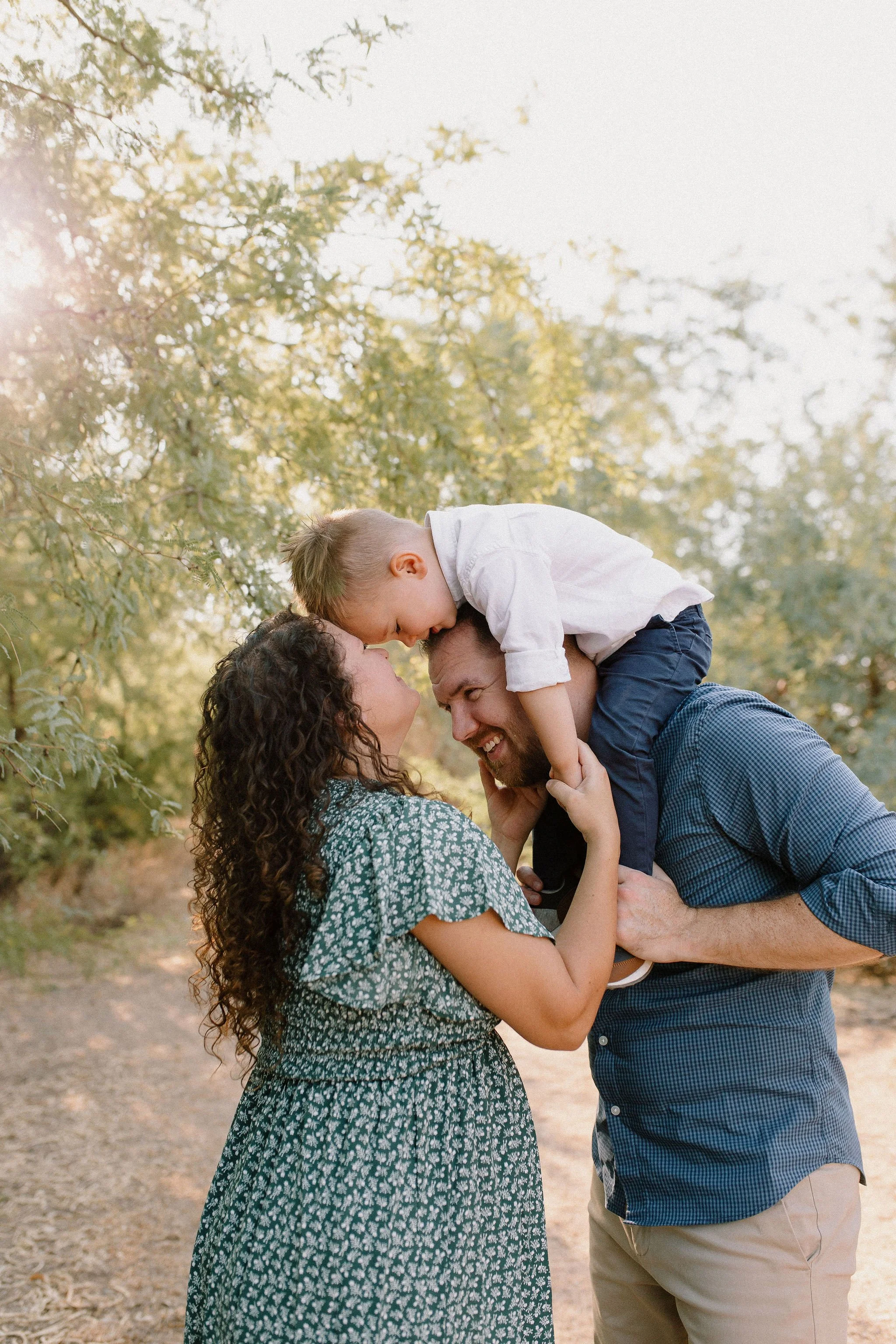 toddler sitting on dad's shoulder leaning down to touch foreheads with mom during family maternity photography session