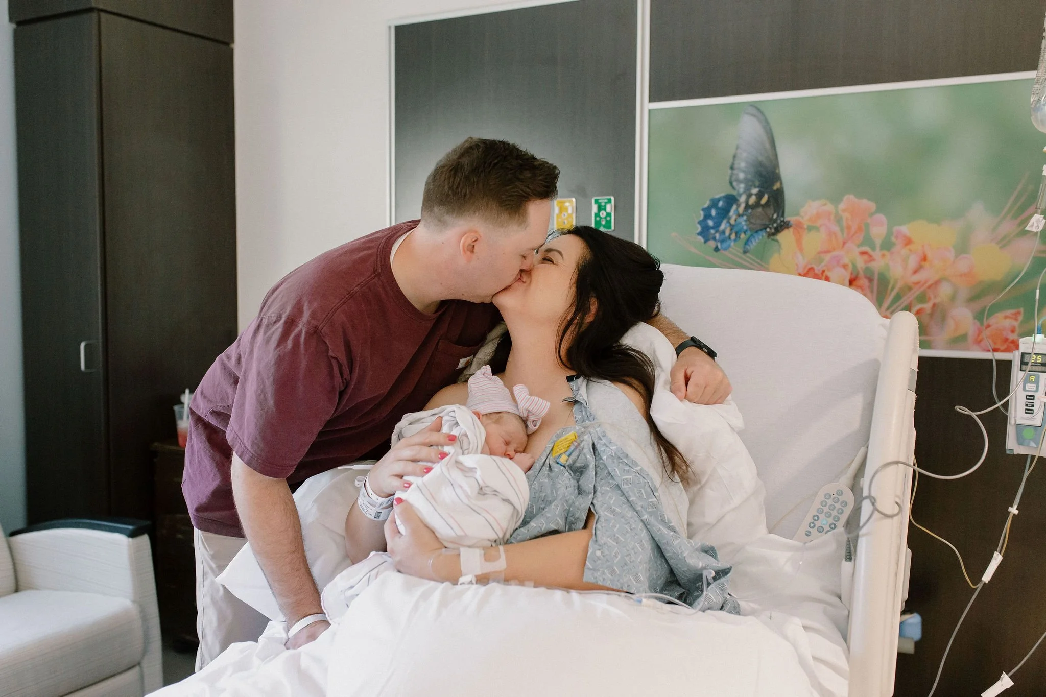 mom and dad kissing while holding newborn baby after birth in hospital birth photography session