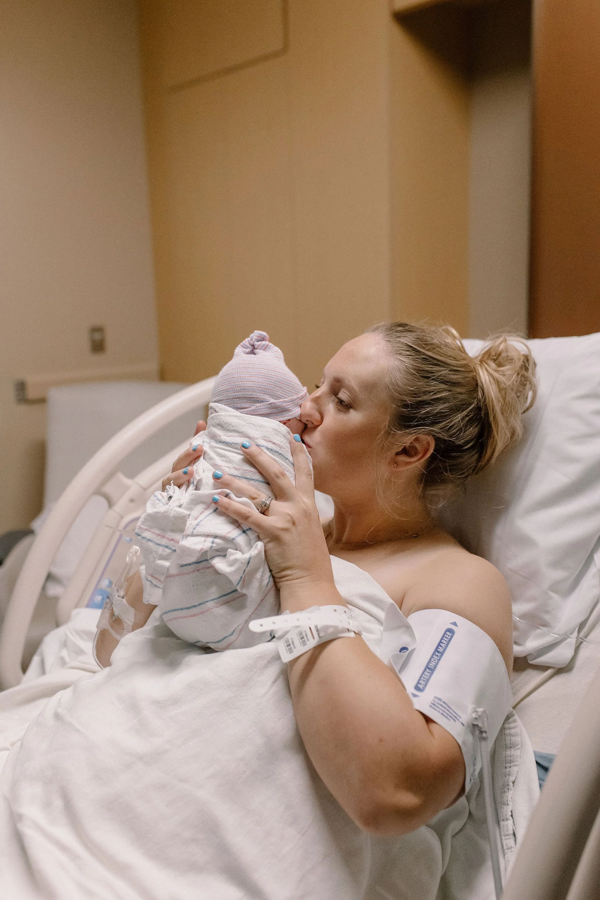 mom kissing swaddled newborn baby in hospital bed after birth