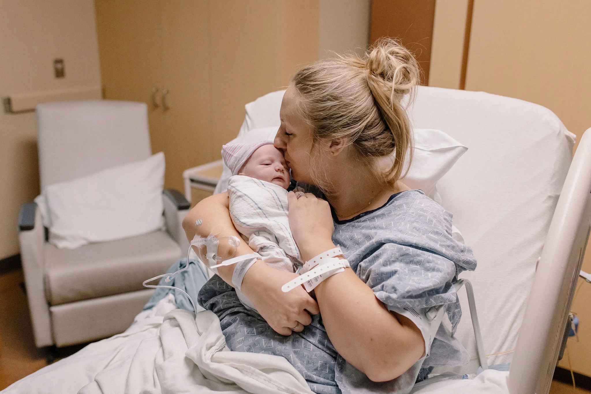 mom kissing swaddled newborn baby in hospital bed after birth