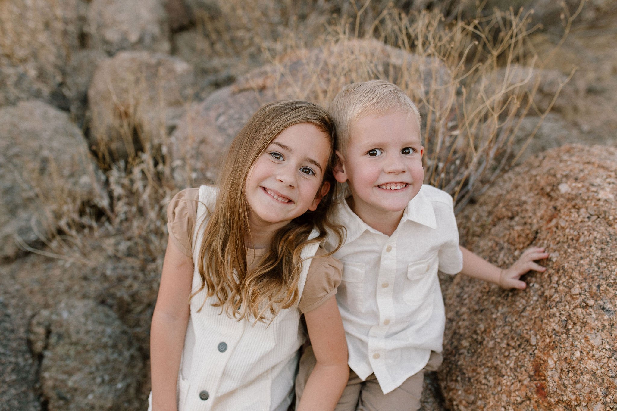 siblings sitting close on boulders smiling during desert family photography session in Scottsdale, Arizona
