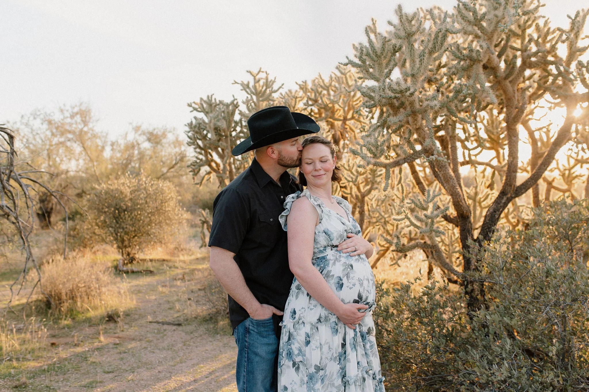 Pregnant mom holding belly husband kissing her head wearing cowboy hat in desert maternity photoshoot in Gilbert Arizona