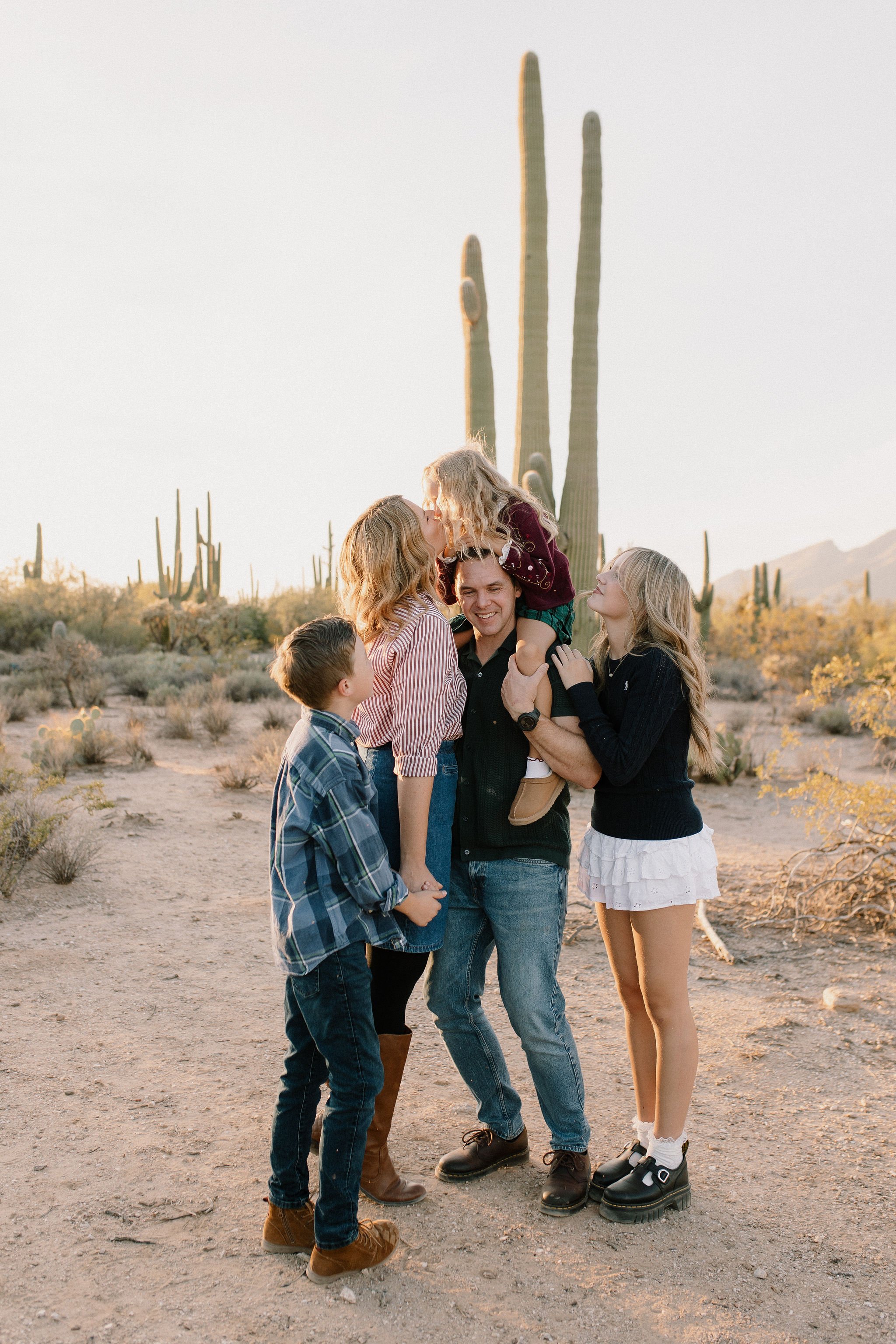 family standing close together looking at mom kissing toddler on dad's shoulders in the desert