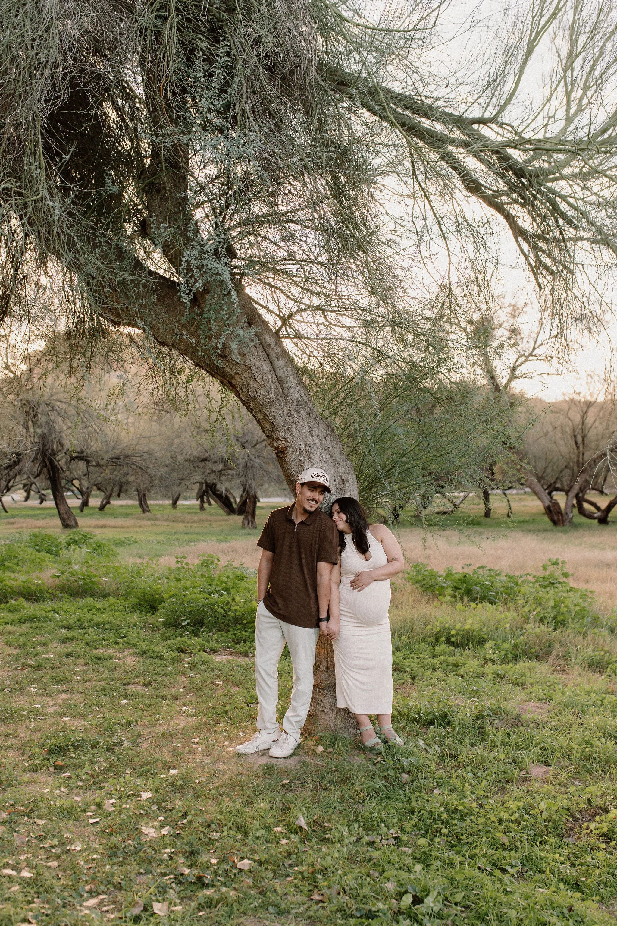 pregnant mom and dad cuddling and holding hands in a green field with trees