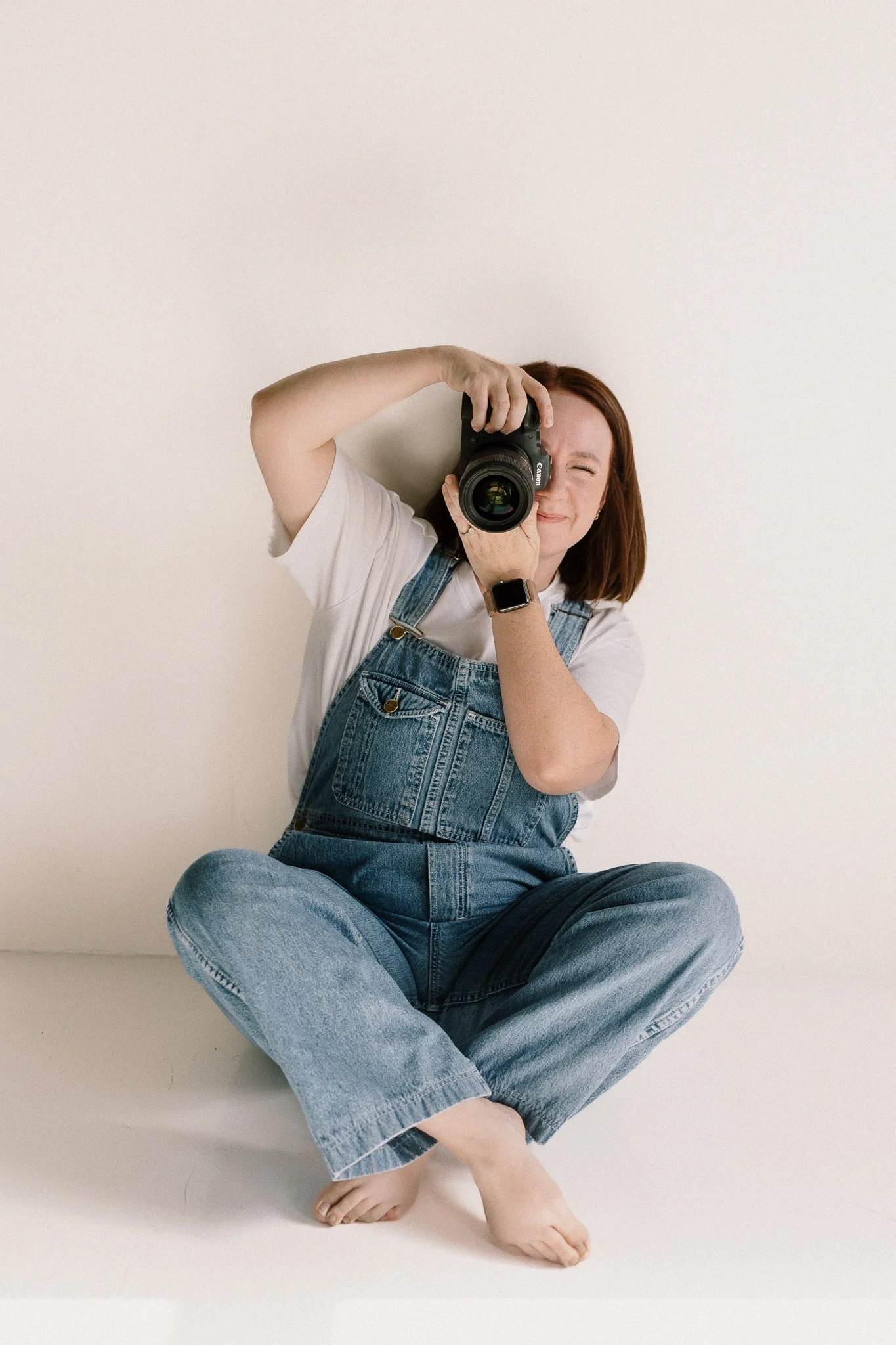 photographer holding camera taking a picture in a neutral studio space in overalls and a white shirt