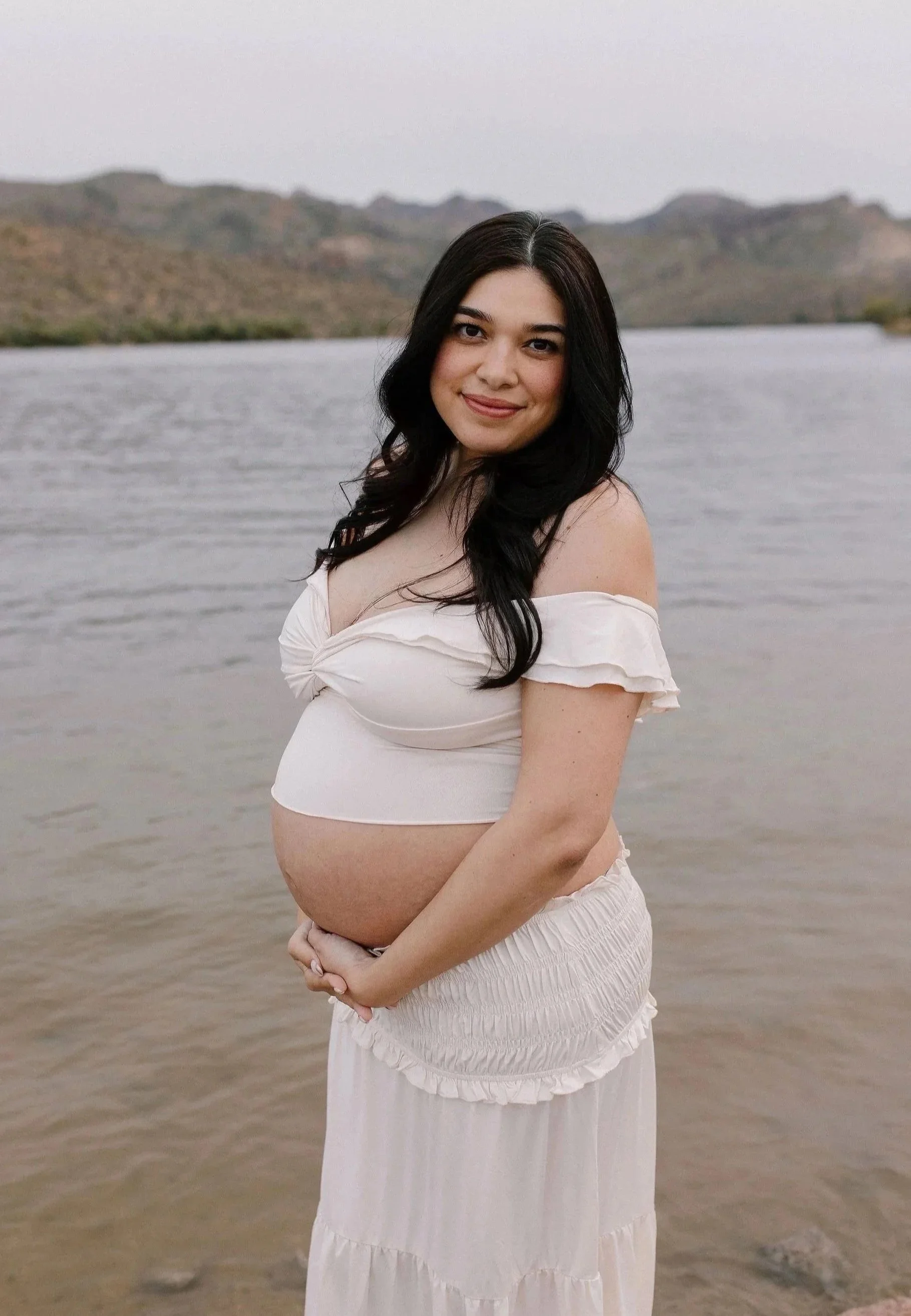 A pregnant woman with dark hair wearing a white off-the-shoulder top and long skirt stands by a body of water with mountains in the background.