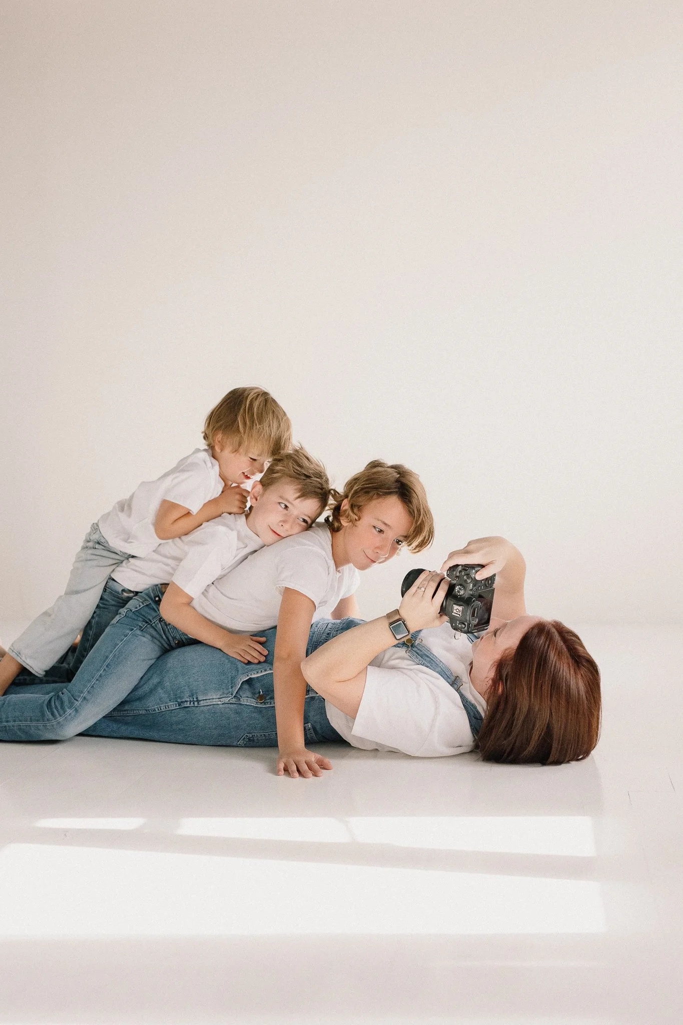 photographer laying on the ground with her children dogpiling her as she takes a photo of them in neutral studio space