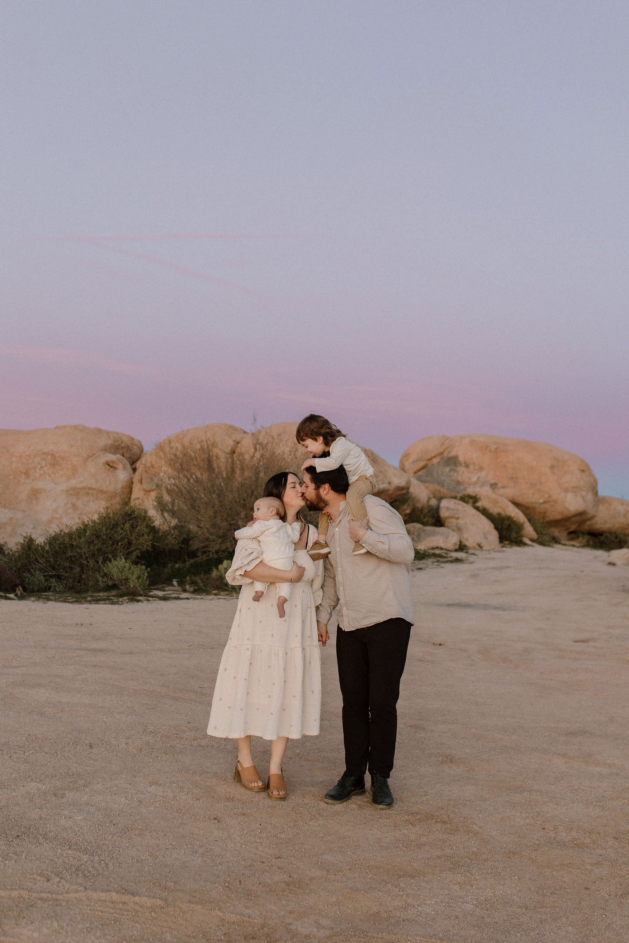 family of 4 walking together in the desert with big boulders mom holding newborn and toddler on dad's shoulders, mom and dad kissing