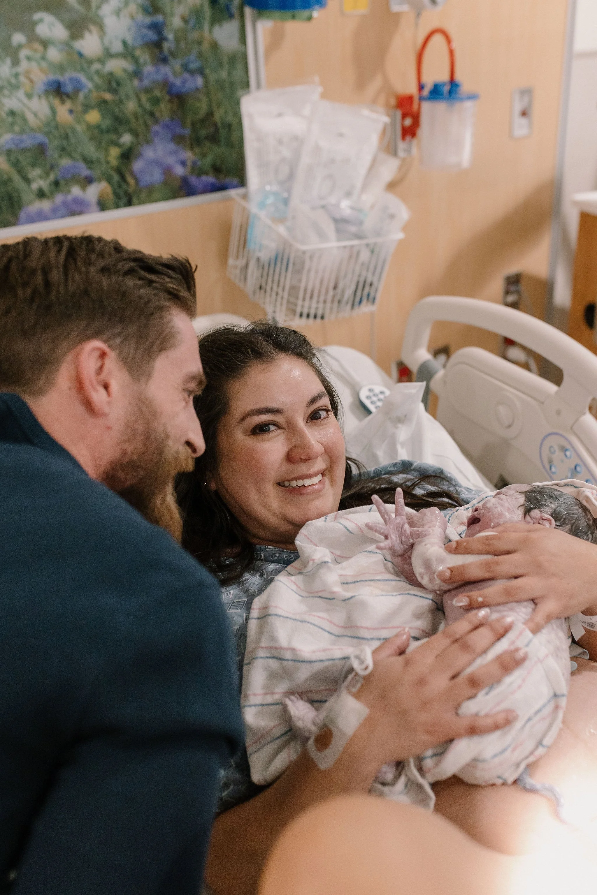 mom holding newborn baby immediately after birth in hospital bed smiling with dad looking at newborn baby