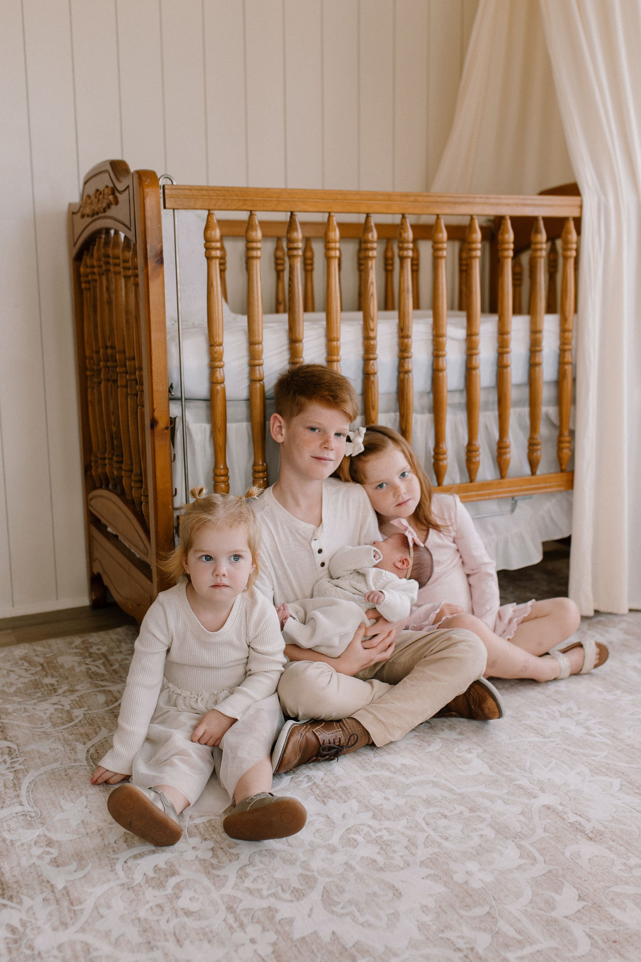 newborn photographer captures siblings holding newborn baby sister in front of crib in lifestyle photography session