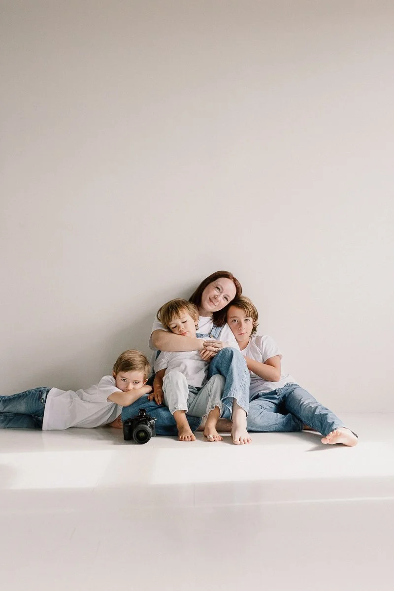 photographer sitting on the ground holding camera with her children sitting close in a neutral studio space