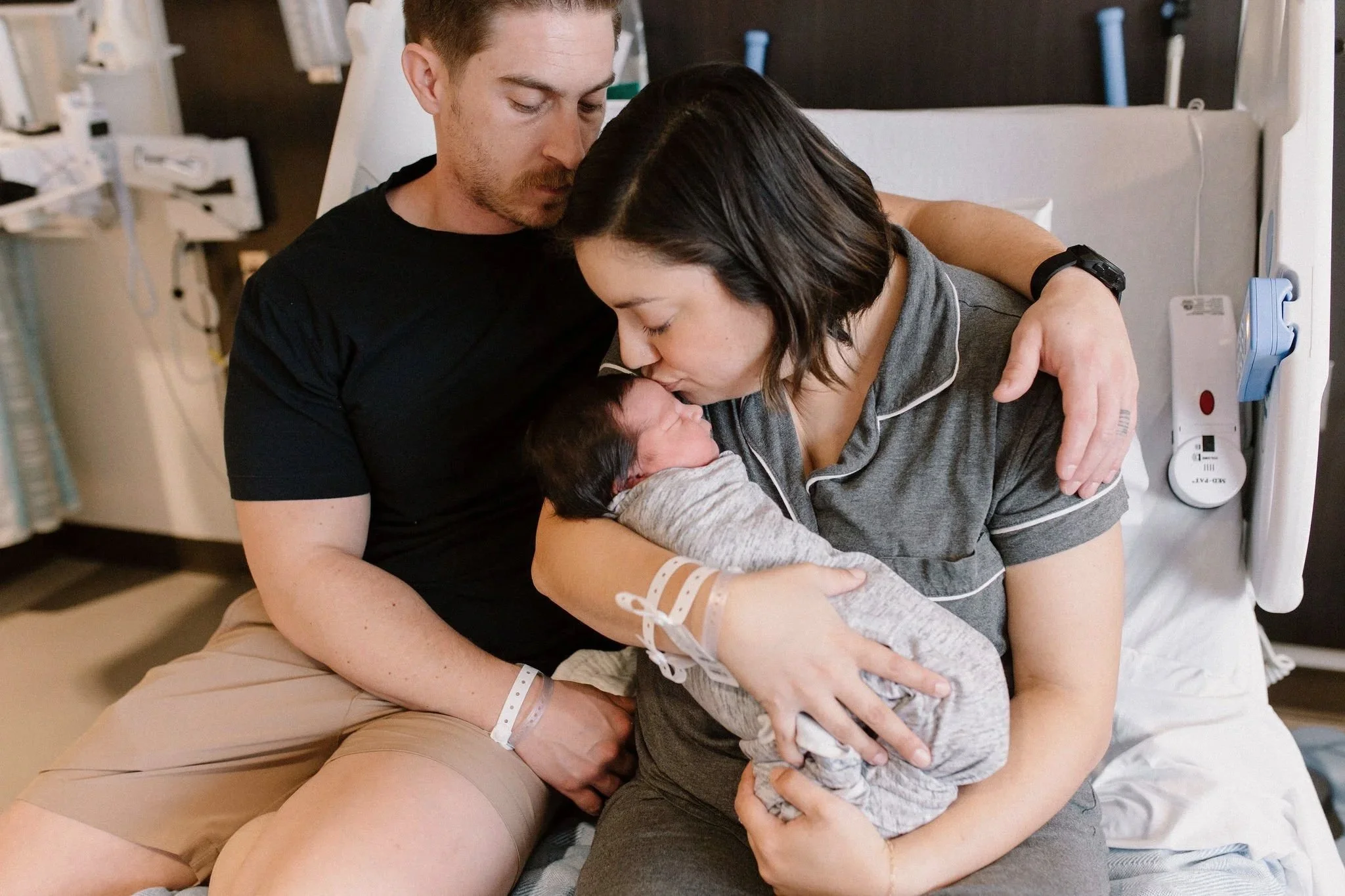 dad holding mom kissing newborn baby in hospital bed captured by Mesa fresh 48 photographer