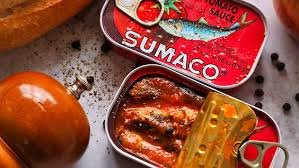 This image depicts two tins of sardines slightly off center in frame. One tin is half peeled open revealing little fillets of sardines packed in tomato sauce. Next to the tins are scattered peppercorns and cracked black pepper and a pepper mill.