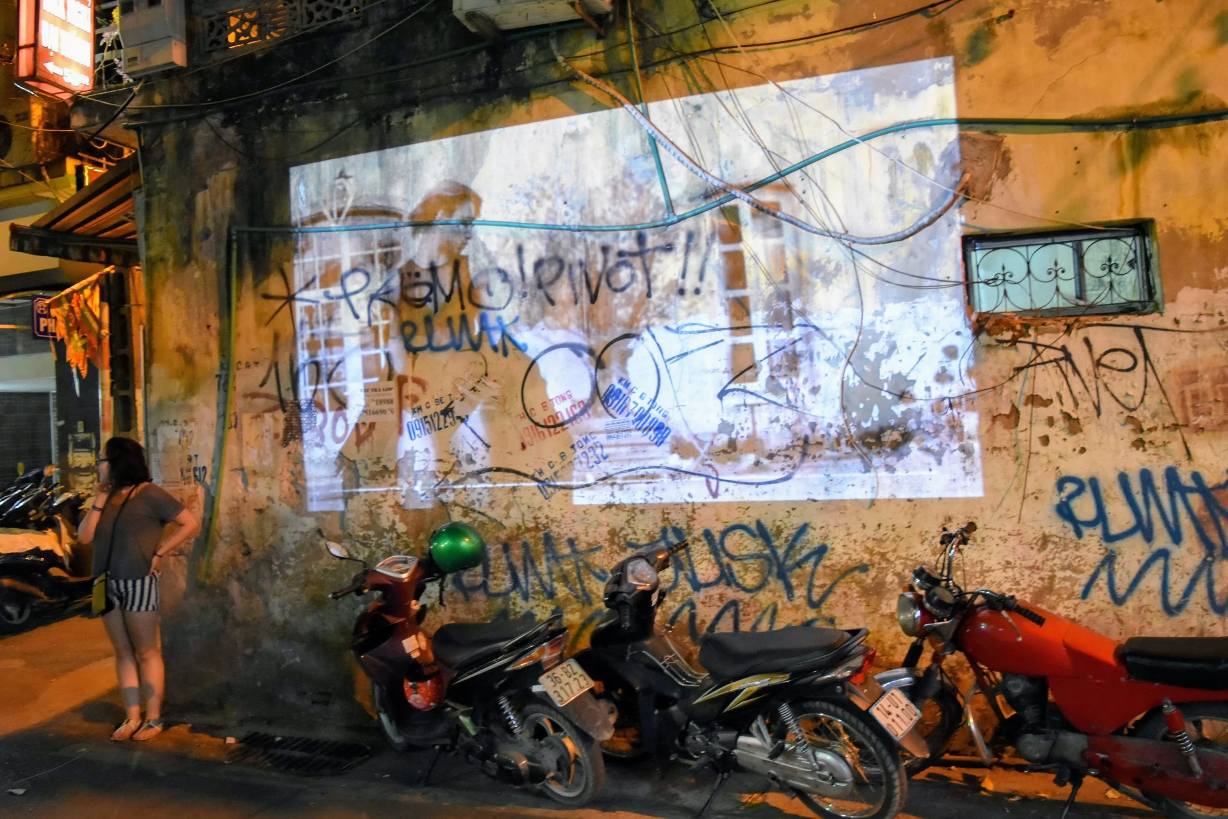 The old quarter in Hanoi. 2 motorbikes parked against a weathered wall with graffiti and a movie being projected on it.