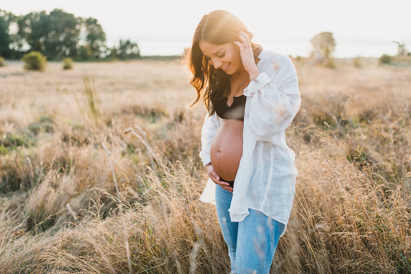 Pregnant woman standing in a grassy field during golden hour, wearing jeans and a white shirt, smiling and holding her belly during a maternity photo session in Seattle.