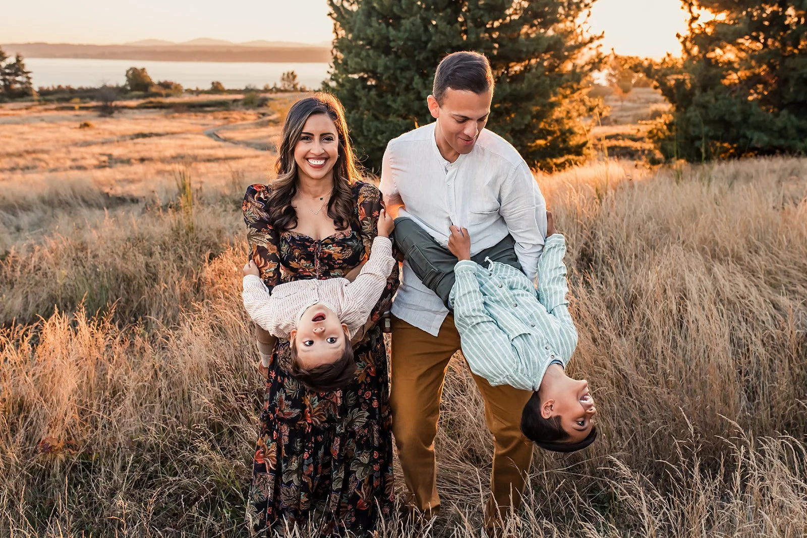 Family playing together in a golden field in Seattle at sunset, with parents holding their kids upside down while everyone laughs during a playful, kid led family photo session.