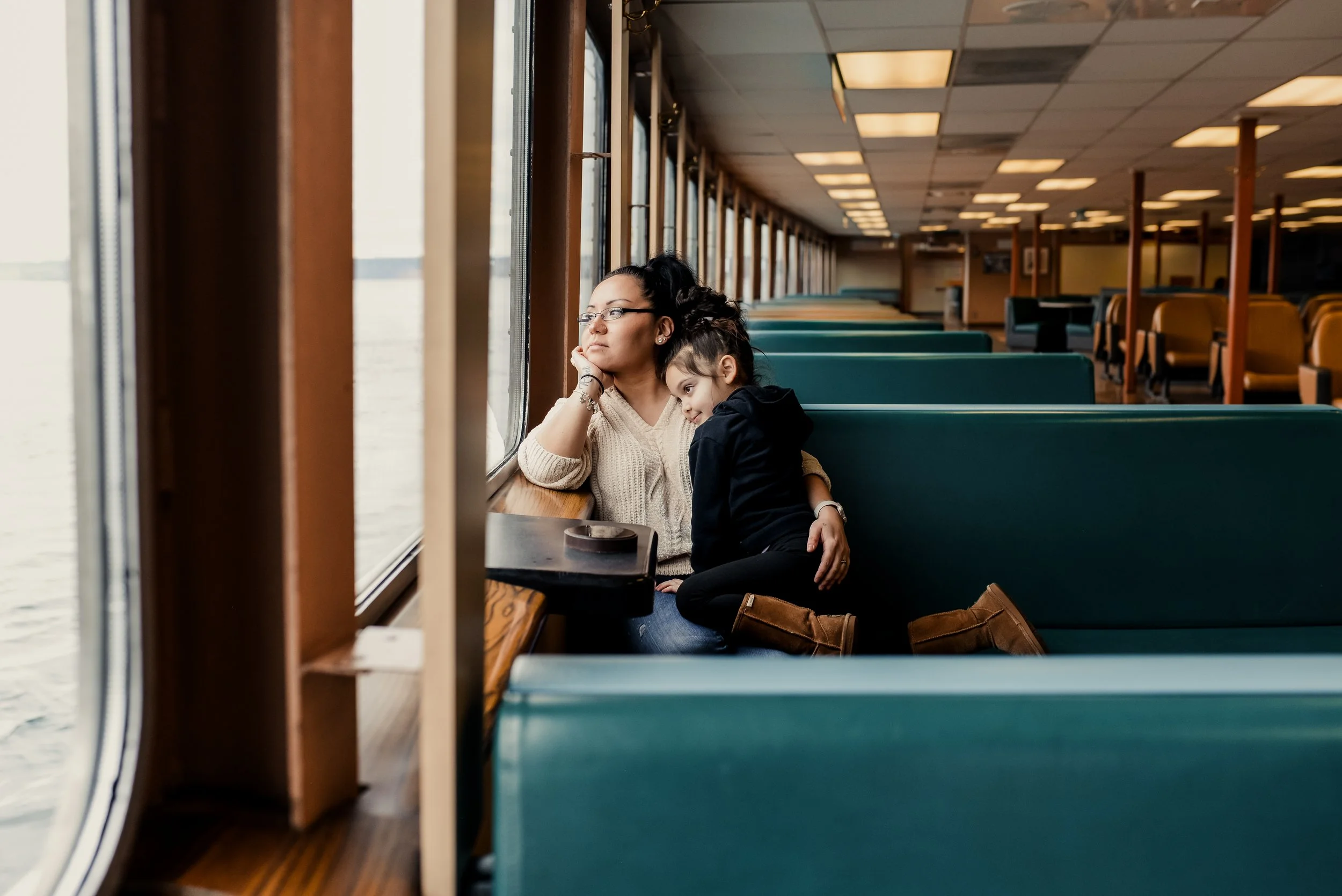Mother and child sitting together on a quiet Washington State ferry, the child resting on her parent’s shoulder while looking out the window during a calm, natural family photo session.