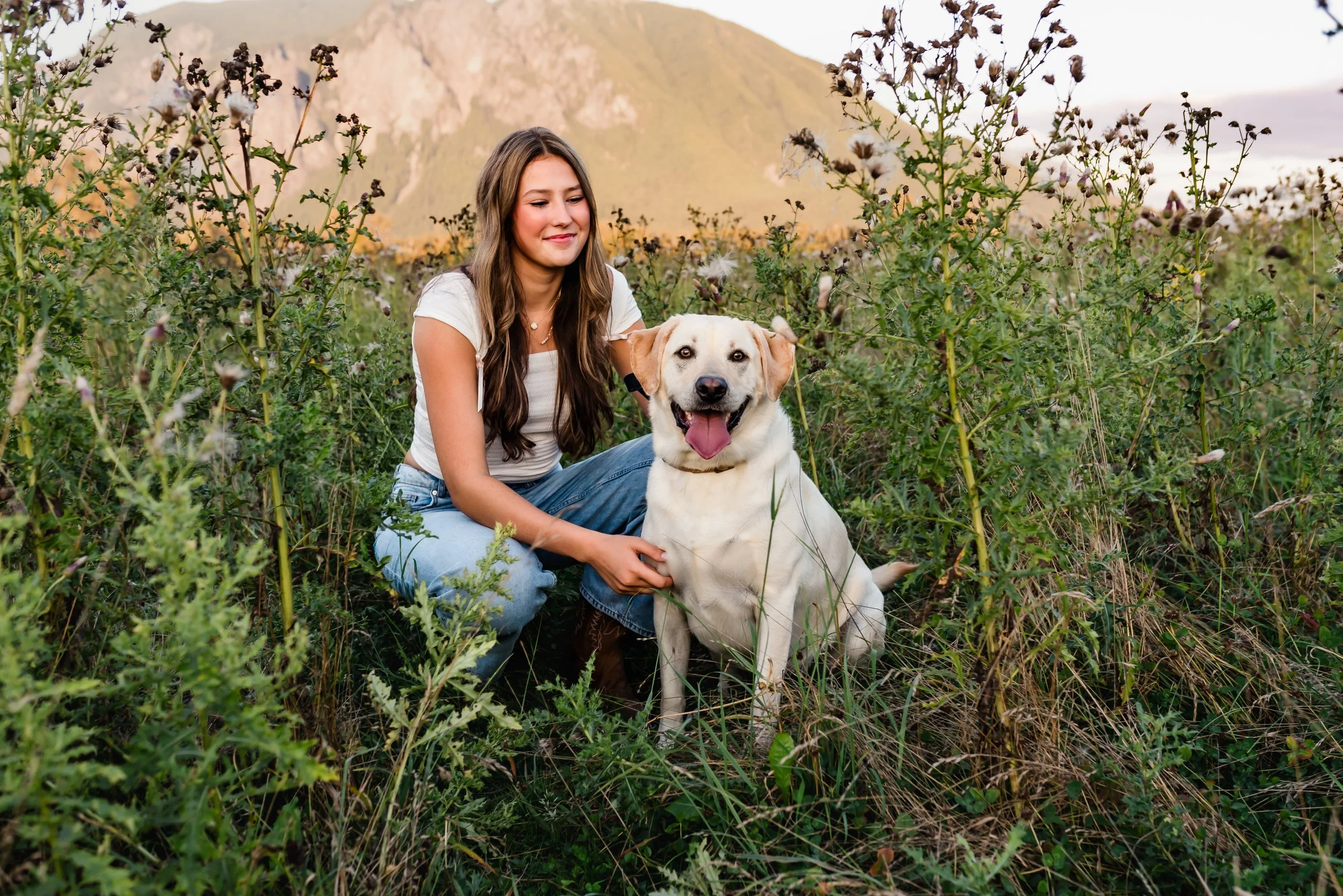 High school senior sitting in a grassy field with her dog, smiling with a mountain in the background during an outdoor senior photo session.